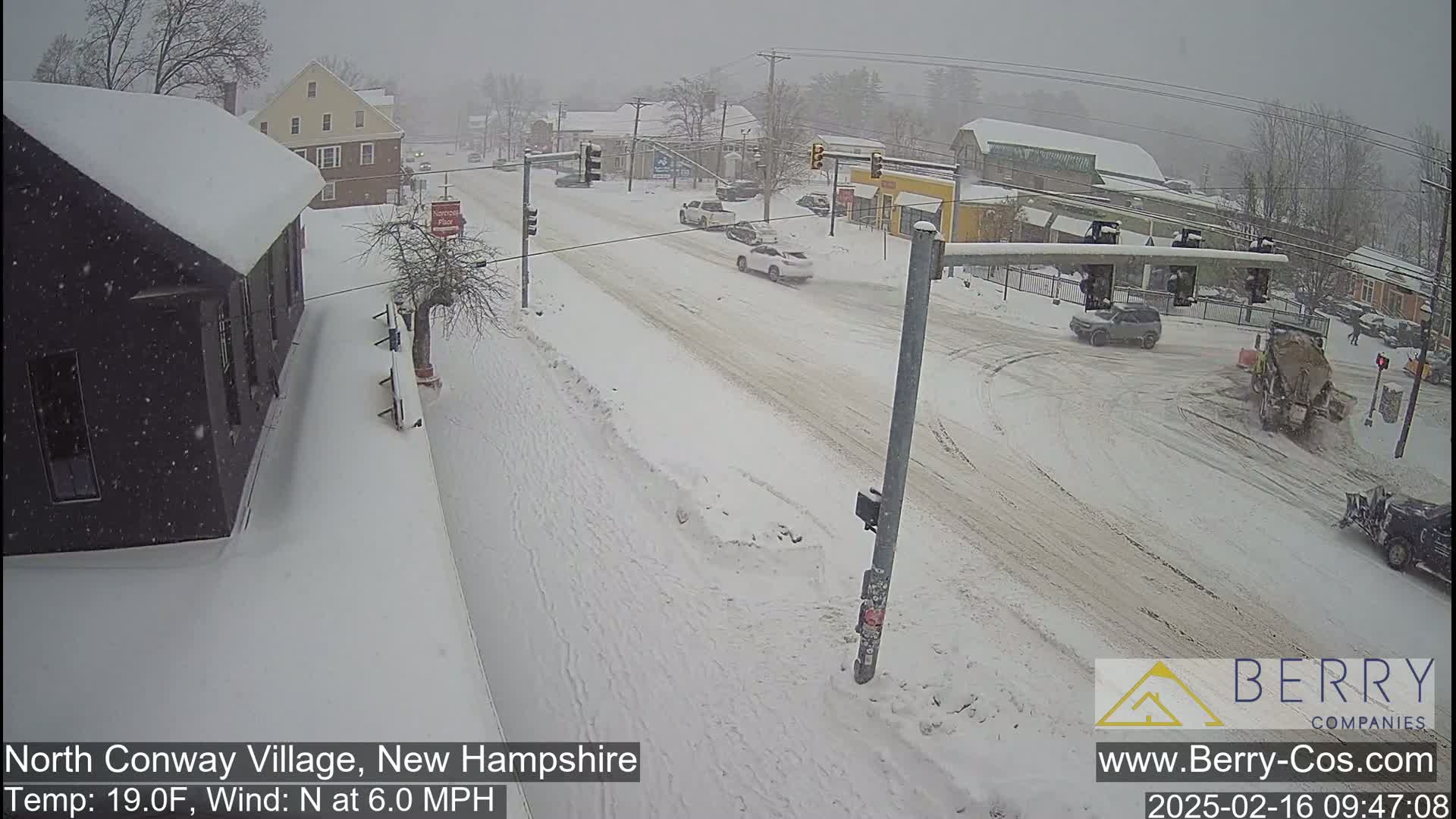 A snow-covered street scene shows several cars navigating a snow-covered intersection during a snowfall, with a snowplow in the background.