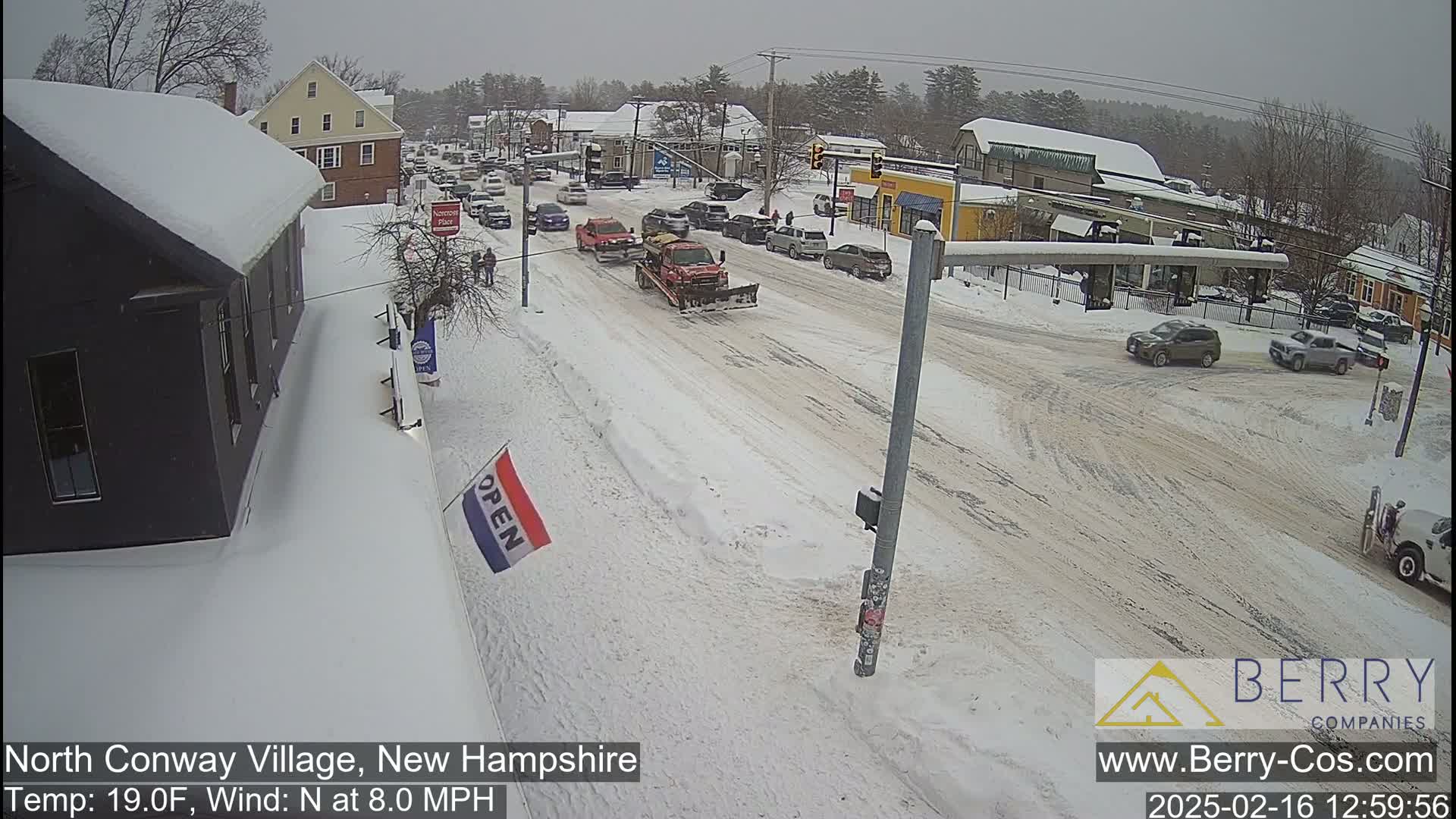 A snow-covered street scene shows several cars and a snowplow amidst buildings under a cloudy sky.