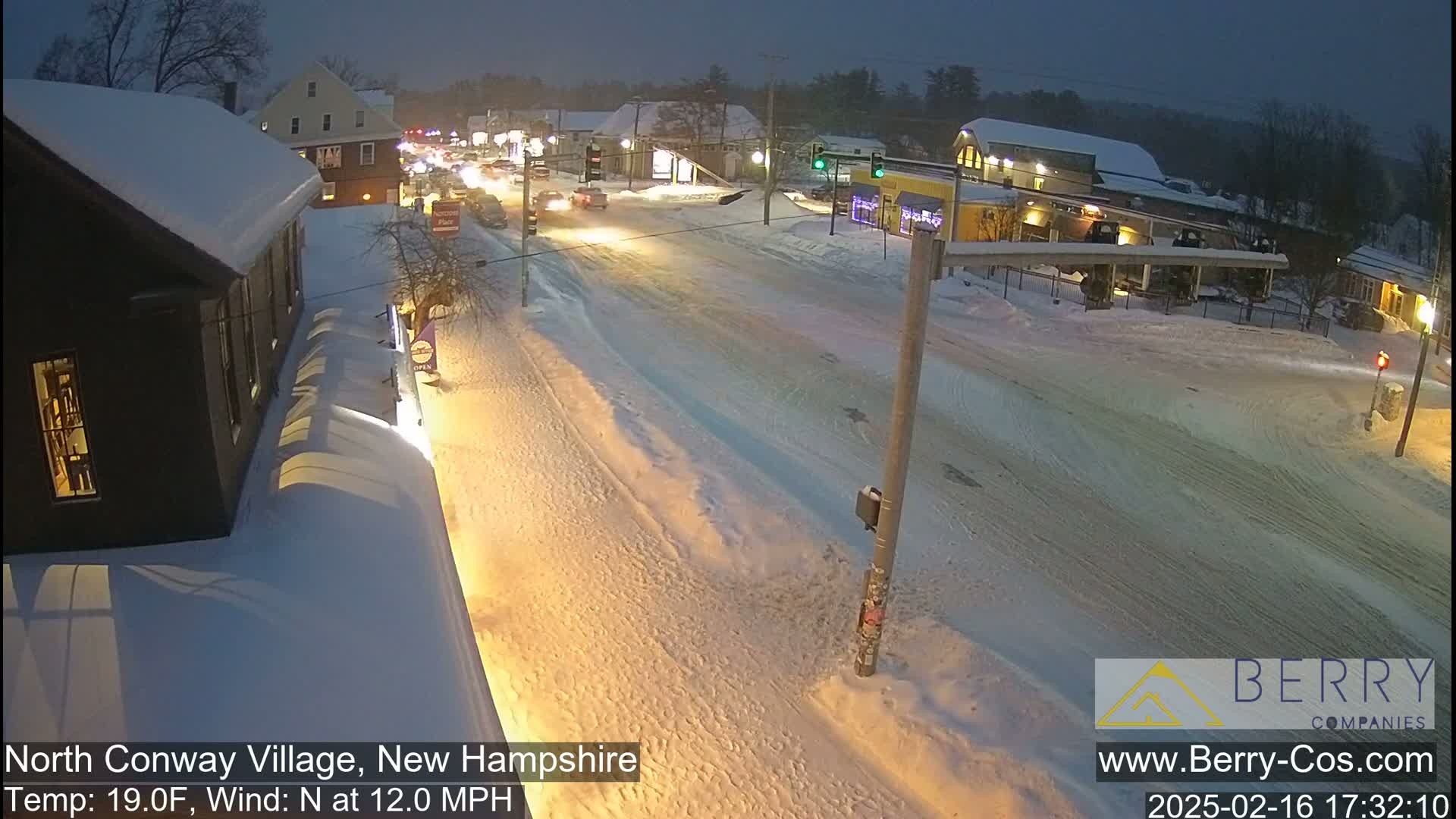 A snow-covered street scene at night, with cars driving on a snow-covered road, under a falling snow and with a temperature of 19 degrees Fahrenheit and a north wind of 12 mph.