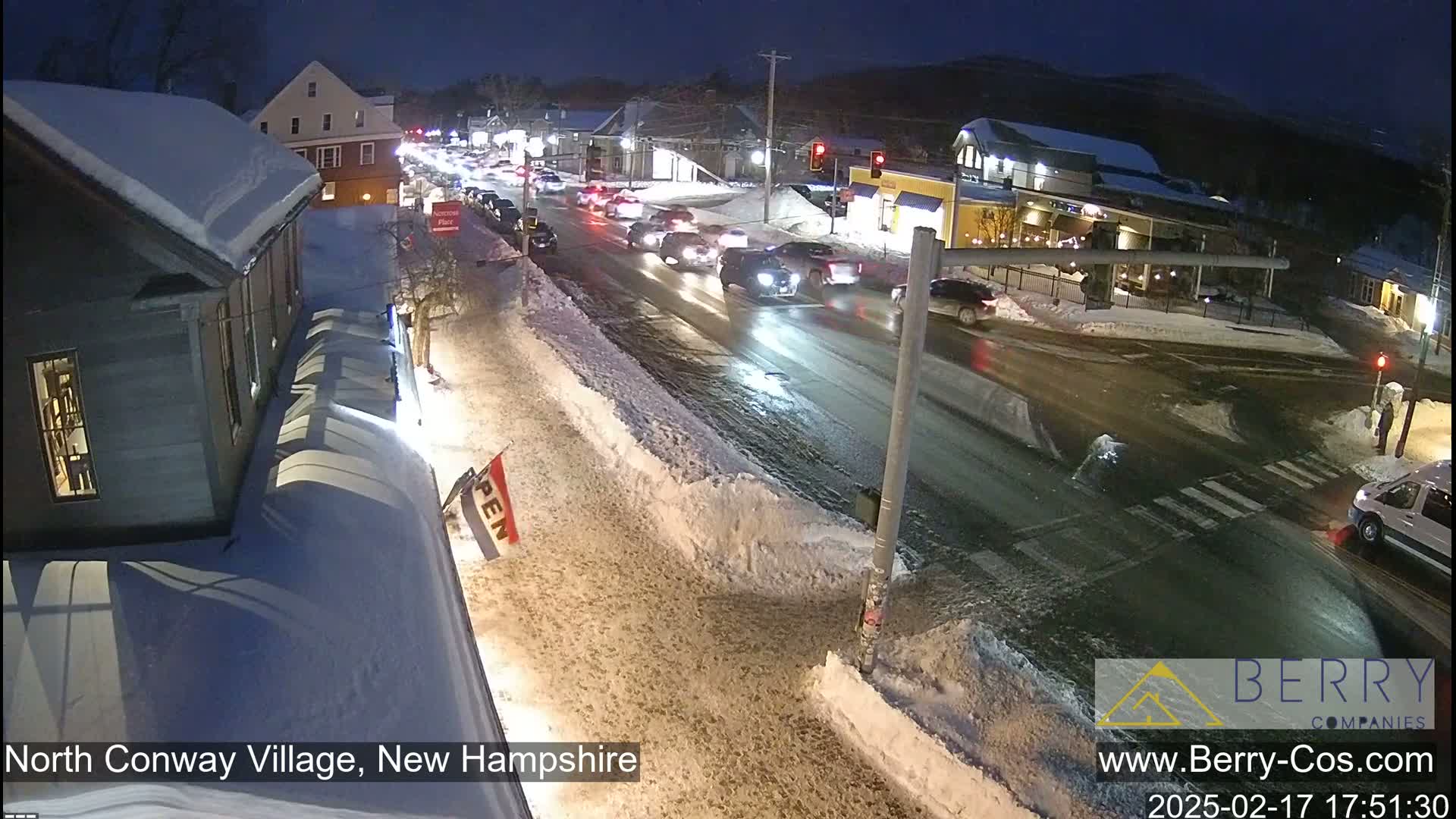 A snow-covered street at night shows traffic moving through a town intersection with several buildings visible under dark, nighttime skies.