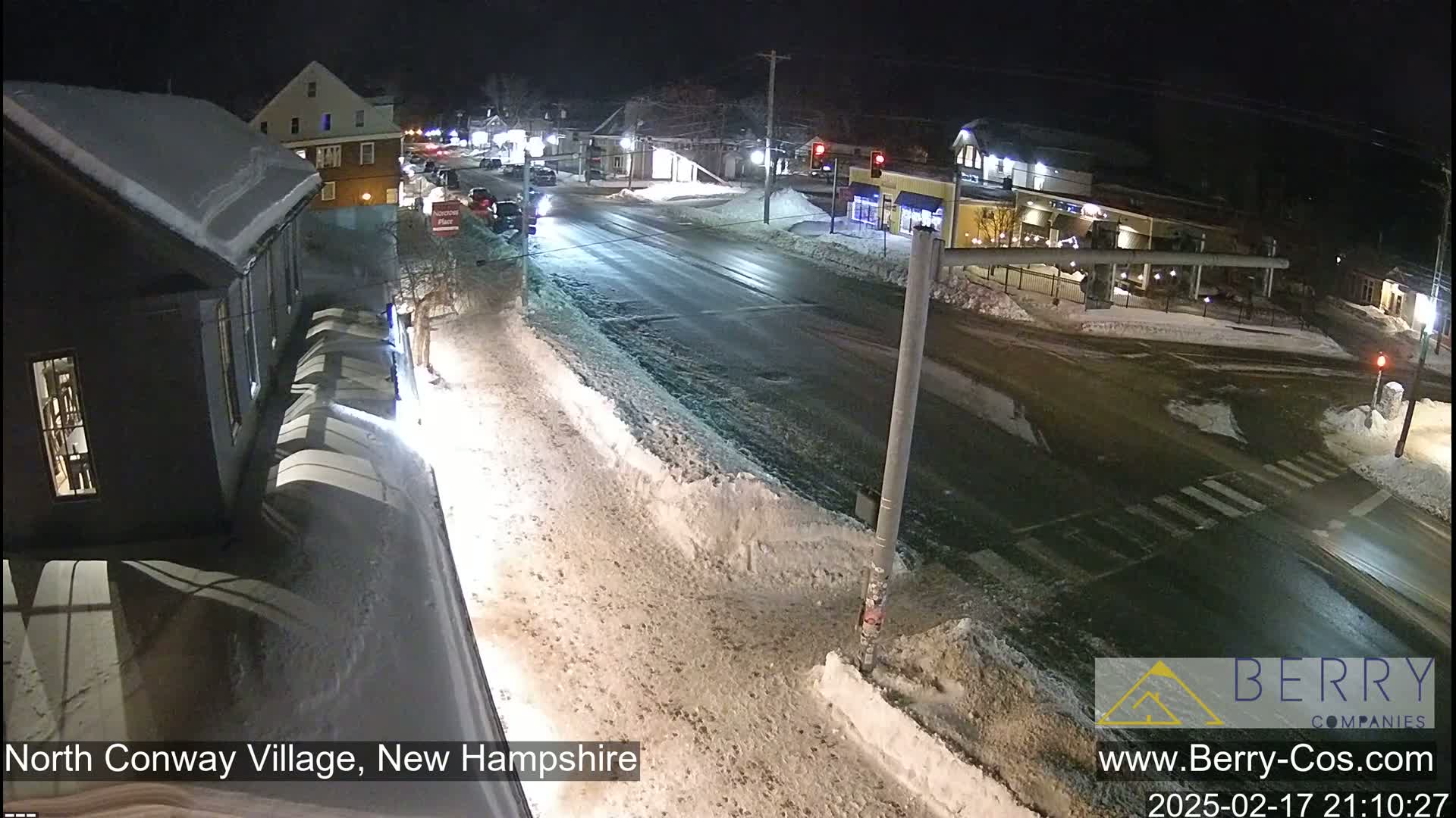 A snow-covered street intersection at night, with buildings and cars visible under streetlights.