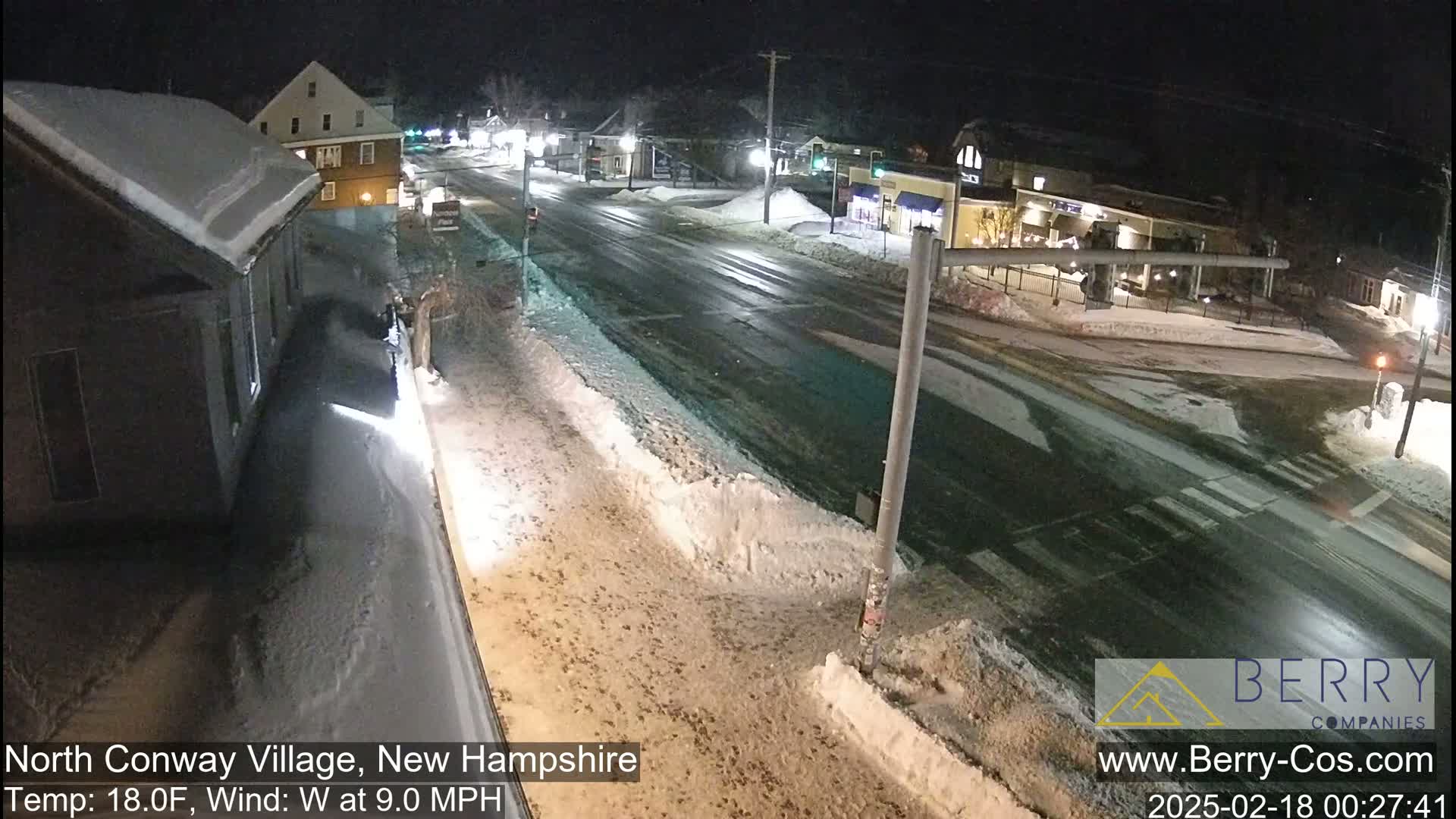 A snow-covered street intersection at night, with buildings and streetlights visible, under a clear sky with a temperature of 18°F and a 9.0 mph west wind.