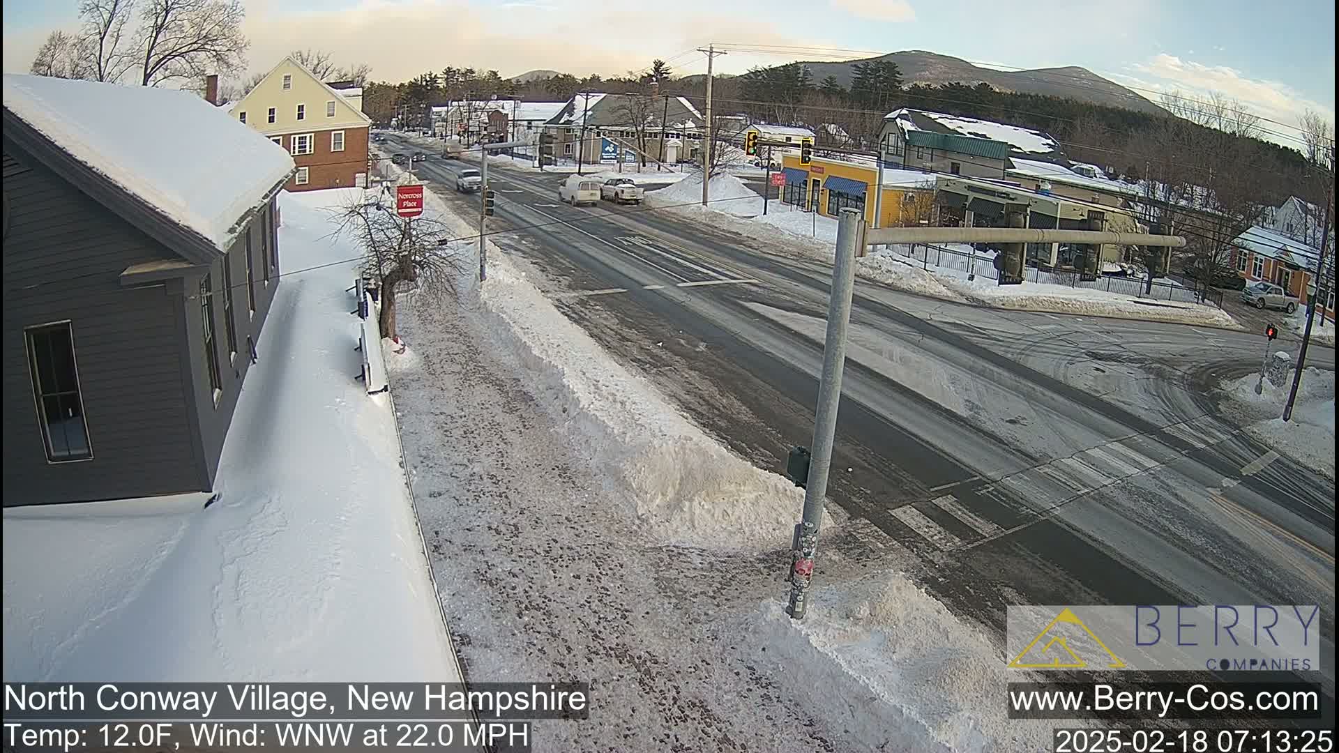 A snow-covered street scene shows a multi-lane road intersection with several vehicles and buildings, under a partly cloudy sky.