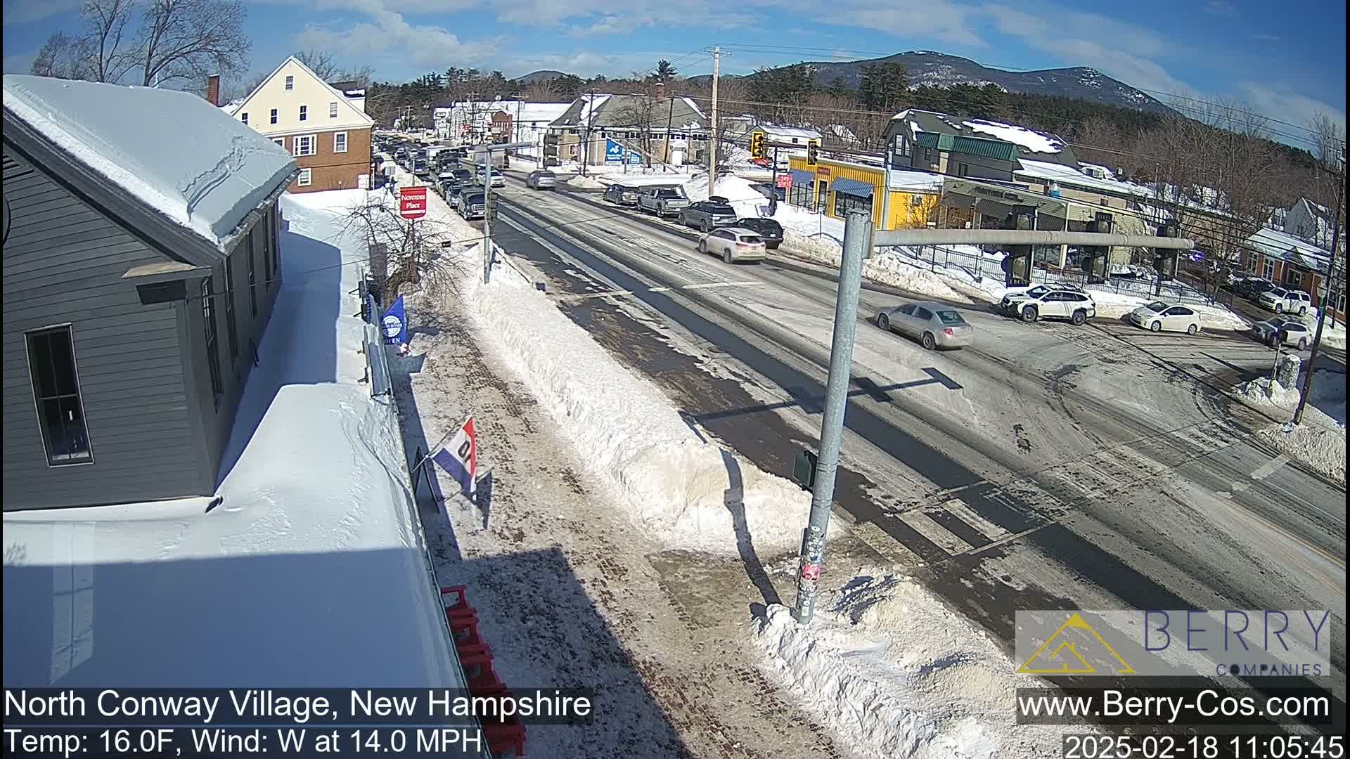 A sunny winter day shows a snow-covered street scene with cars driving on a road lined with buildings, some snowbanks, and shadows cast by the buildings and the sun.