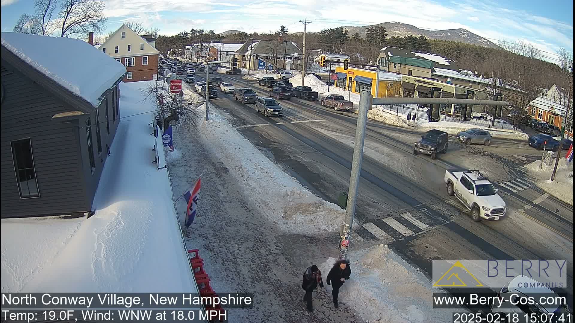 A snow-covered street scene shows cars driving on a road that intersects with another, with pedestrians walking on a snow-covered sidewalk, under a partly sunny sky.