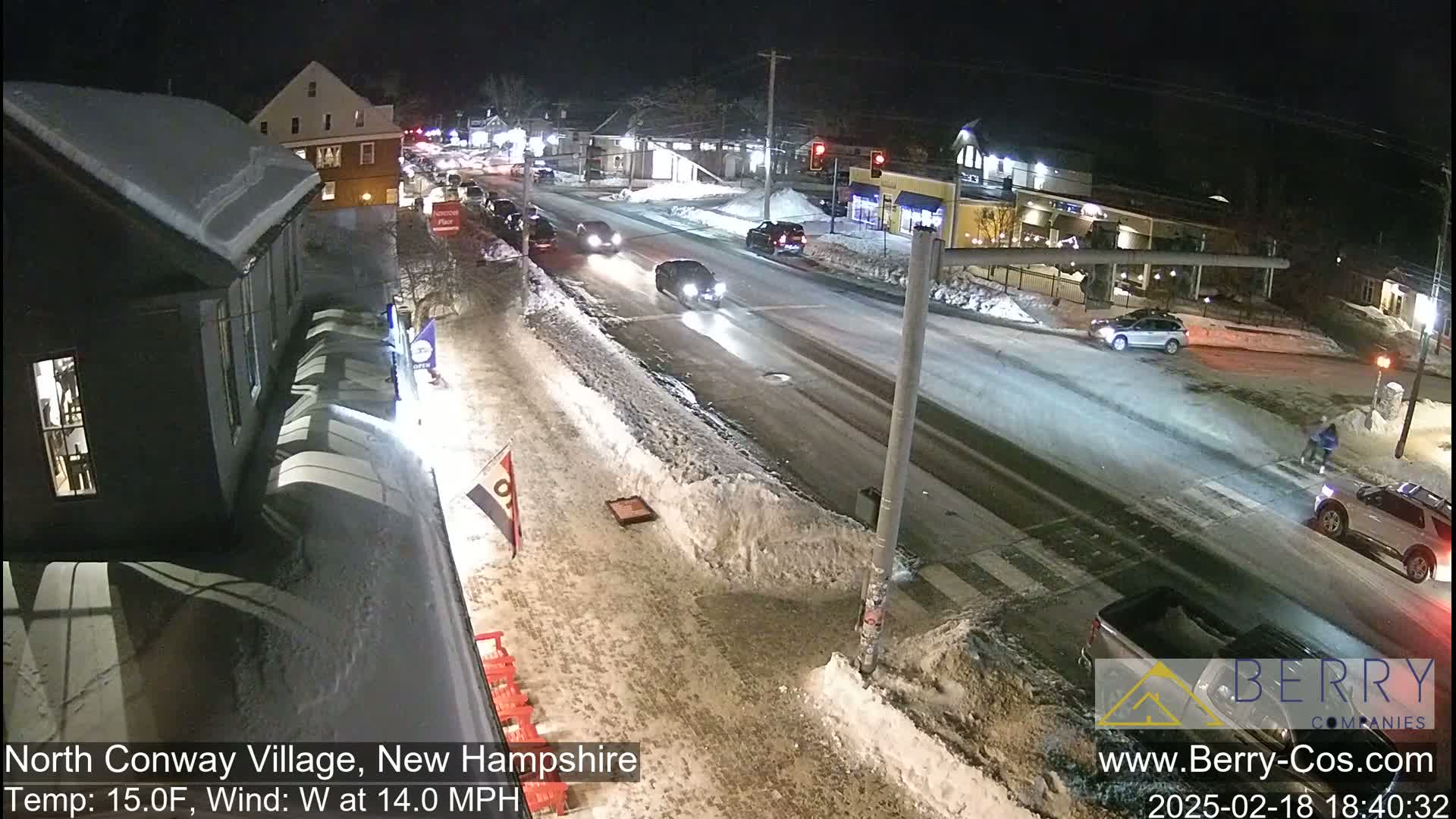 A snow-covered street at night with several cars driving and pedestrians crossing, under a 15°F temperature and 14 mph west wind.