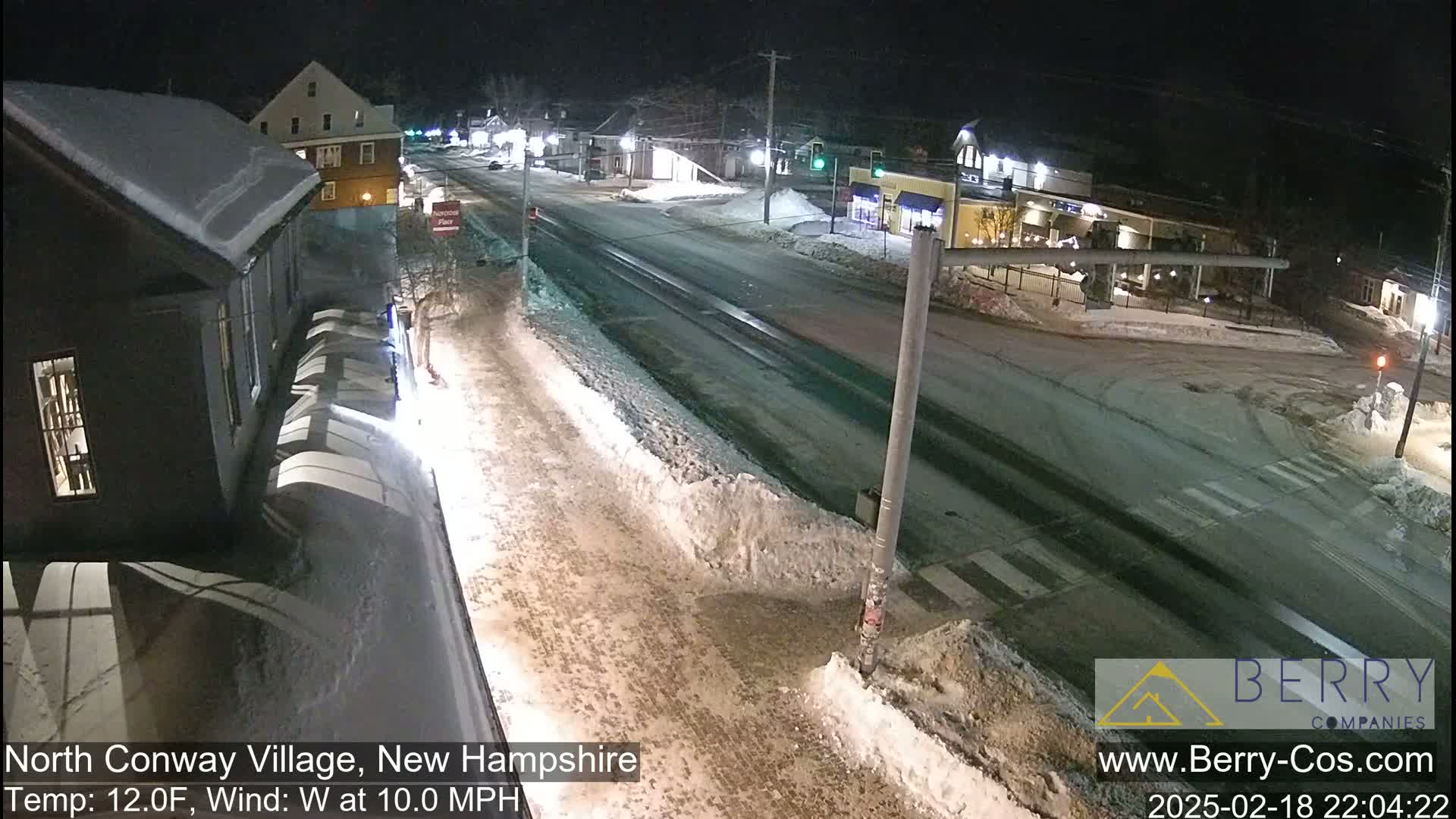 A snow-covered street scene at night, showing a road intersection with buildings on either side, under a dark sky with a 10 mph westerly wind and a temperature of 12°F.