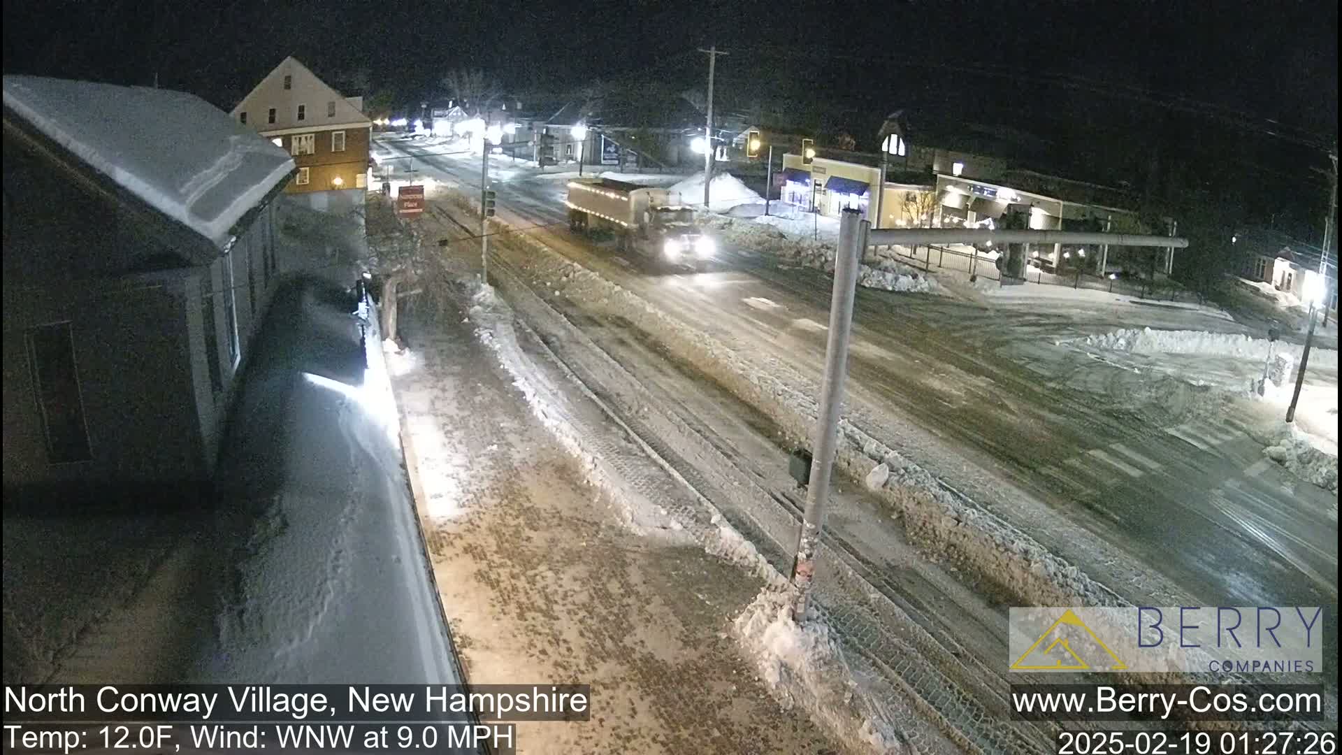 A snow-covered street scene at night, with a large truck driving on a slushy road, under a 12°F temperature with a west-northwest wind at 9 mph.