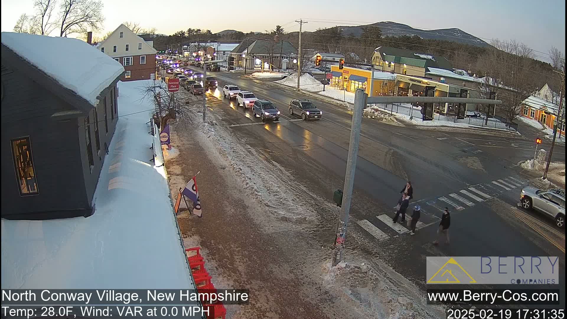 A snow-covered town street scene at dusk shows several cars driving on a wet road with pedestrians crossing a crosswalk, under a clear sky.