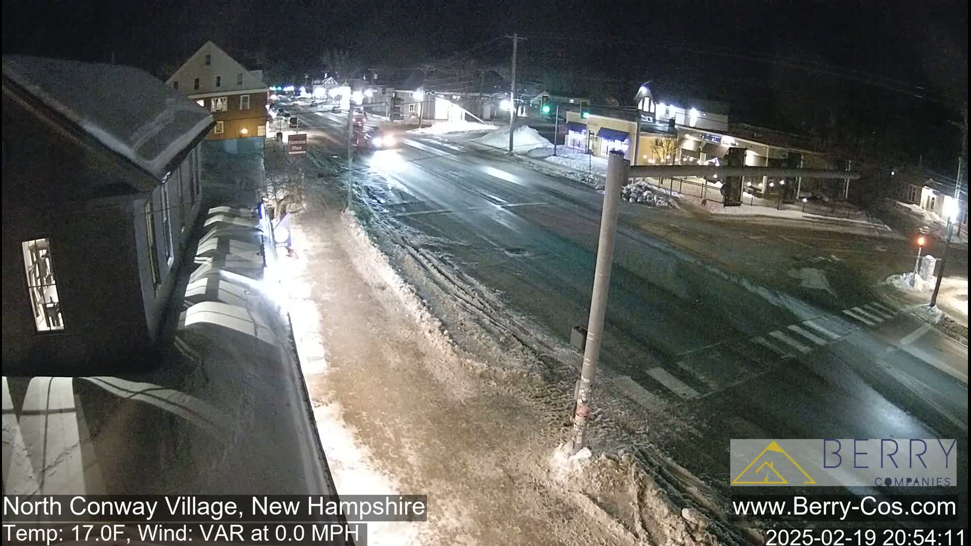A nighttime view of a snow-covered street intersection in a small town, with a few vehicles visible and calm weather conditions.