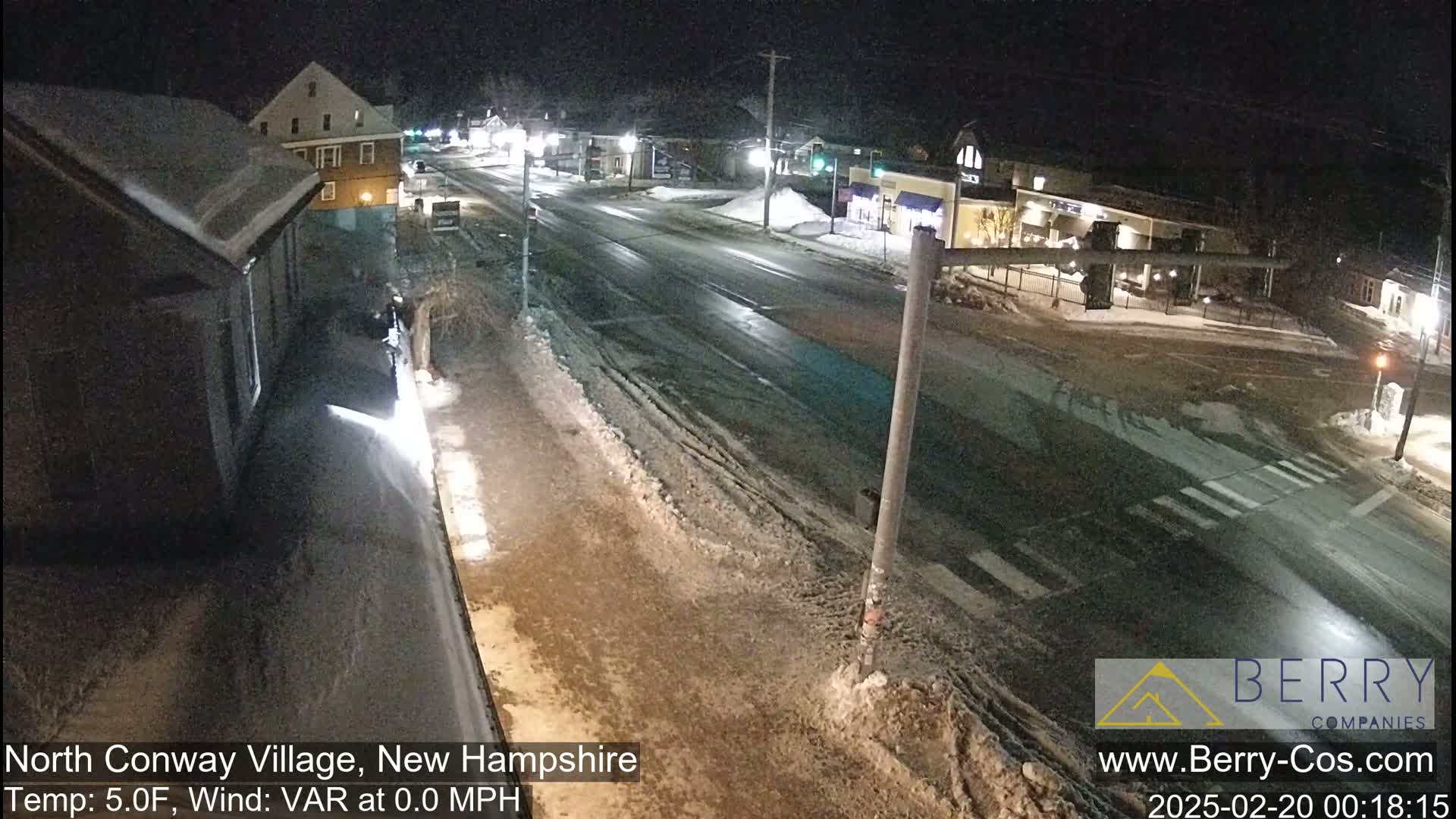 A snow-covered street scene at night shows a multi-lane road intersecting with another road, surrounded by buildings under a dark sky with a temperature of 5°F and a variable wind of 0 mph.