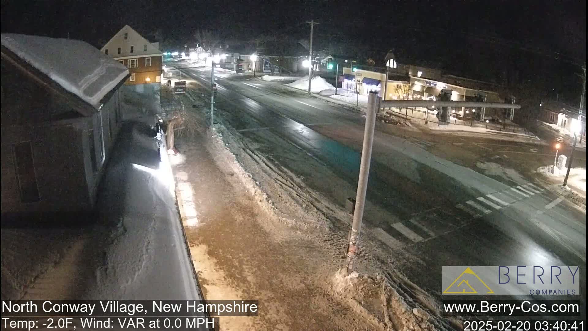 A snow-covered street scene at night shows a mostly empty road with buildings along the sides, under a dark sky.