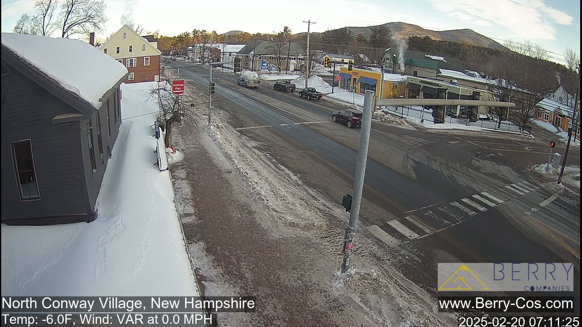 A snowy street scene shows a road intersection with several vehicles, buildings, and snow-covered sidewalks under a clear sky.