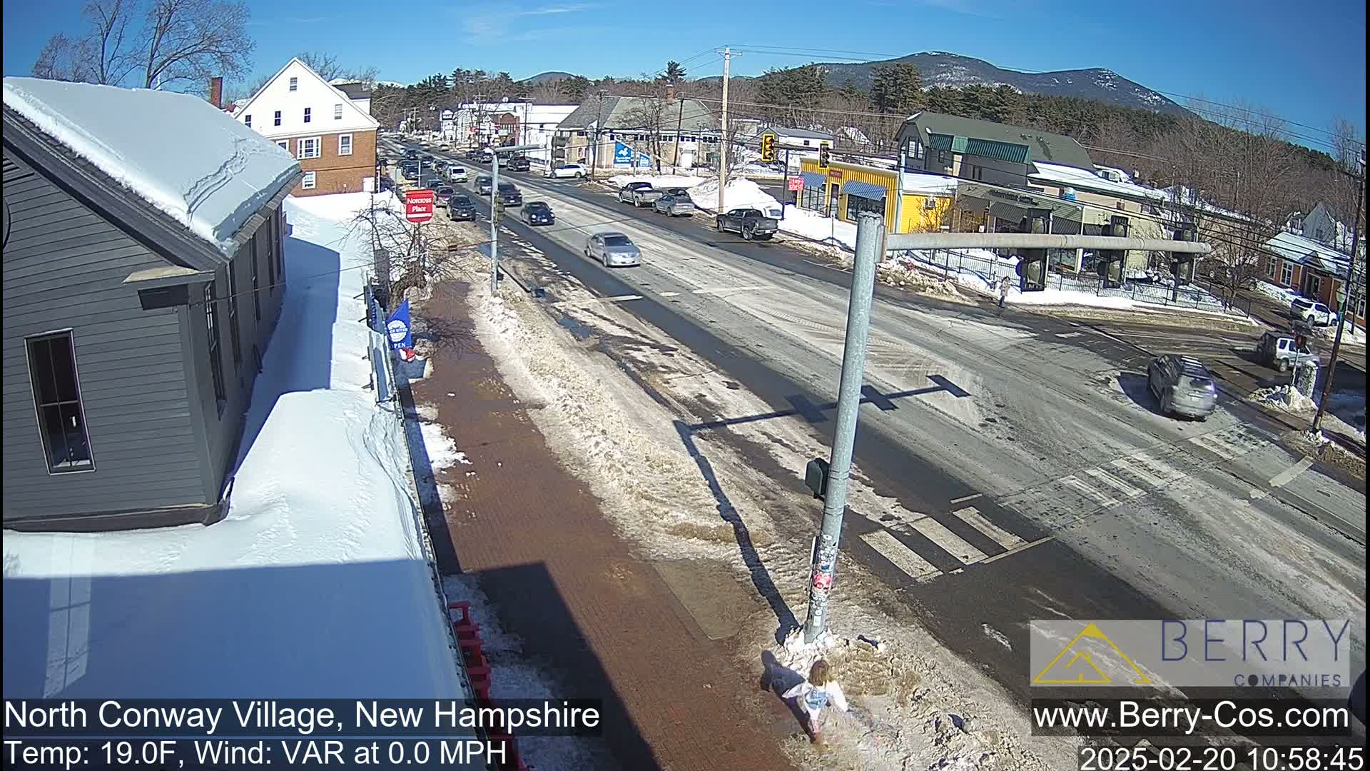 A sunny day in a snow-covered town shows a street intersection with several cars and a pedestrian; melting snow is visible on the road.