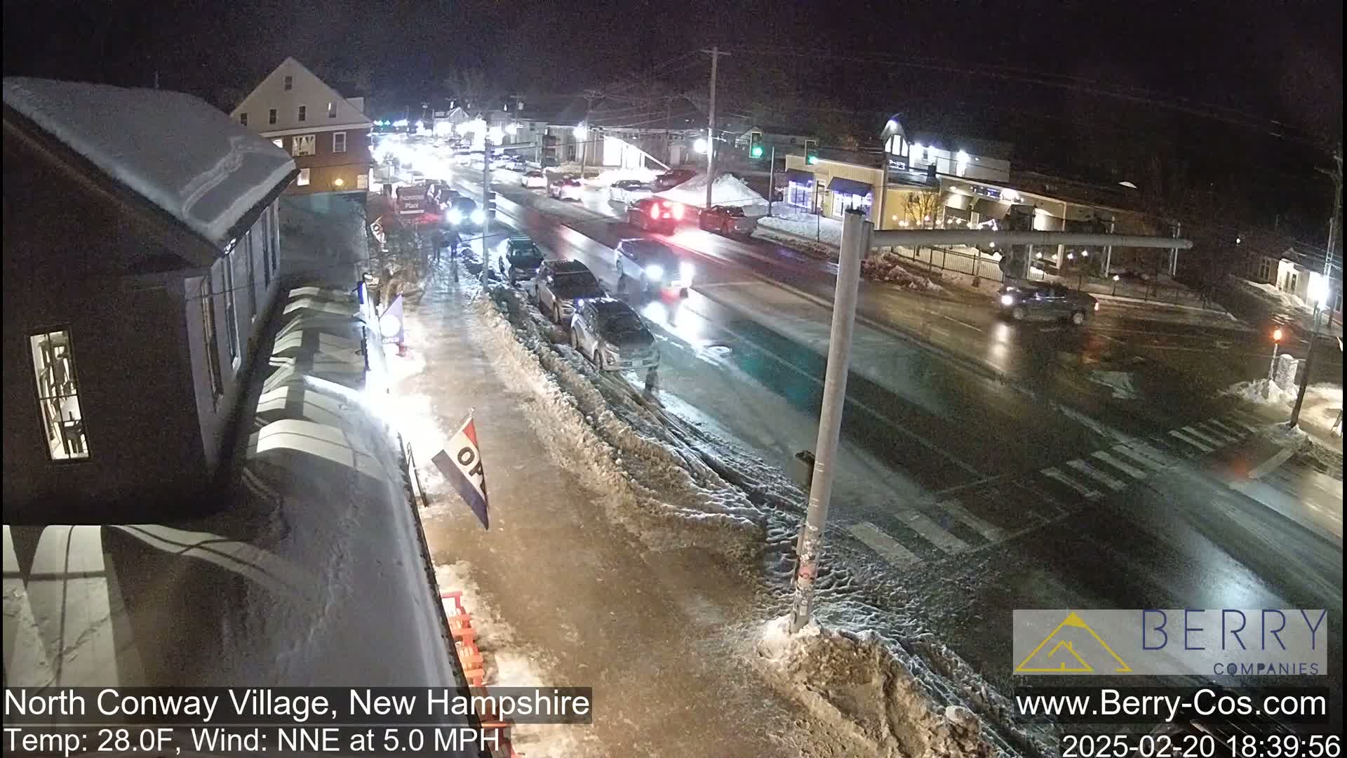 A nighttime street scene shows several cars driving on a snow-covered road, with buildings and snow-covered sidewalks lining the street under partly cloudy conditions.