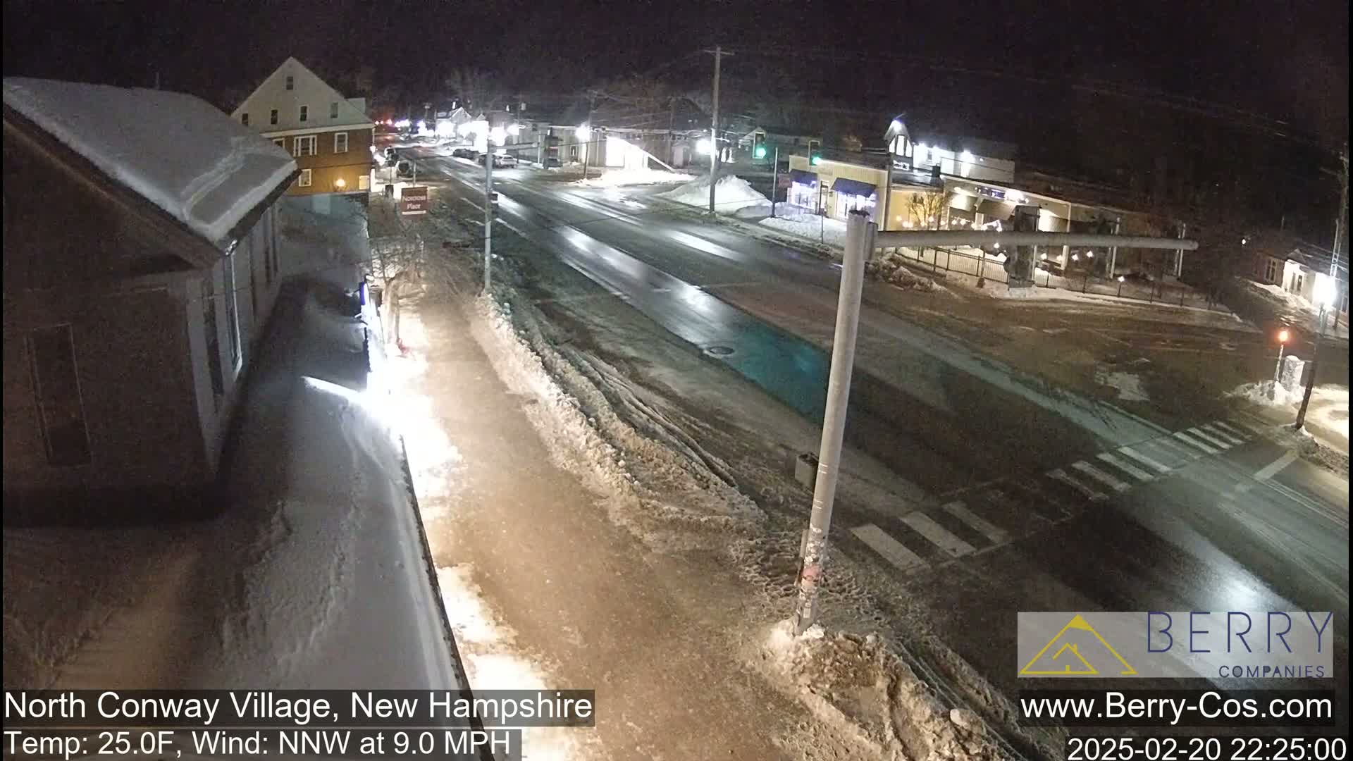A snow-covered street scene at night shows a mostly empty road with buildings and some cars, under a dark sky with a 25°F temperature and a 9 mph north-northwest wind.