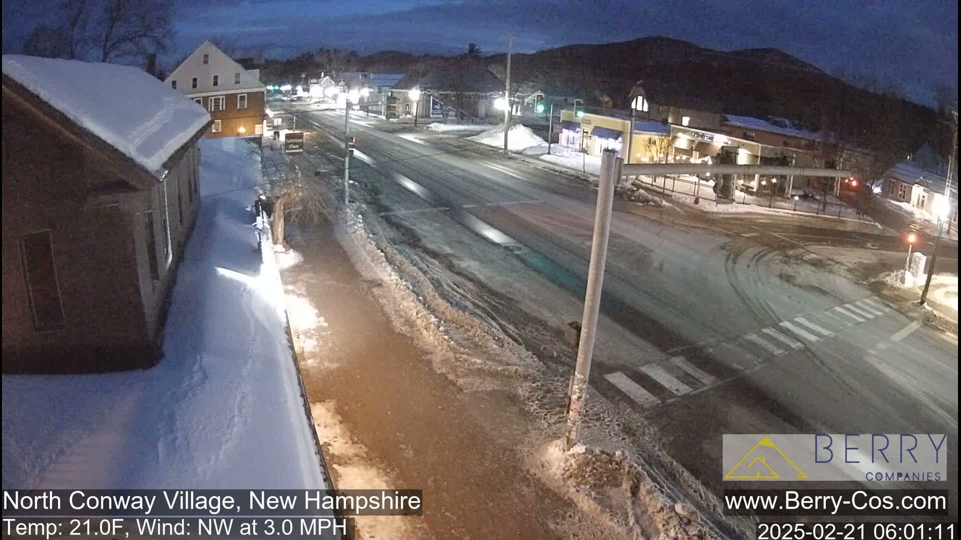 A snow-covered street scene at night shows a quiet town with buildings and roads, under a partly cloudy sky.