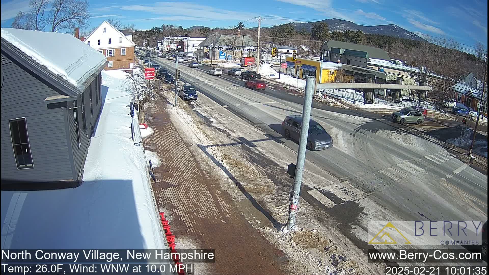 A sunny, snowy street scene shows cars driving on a road that intersects with another, with snow-covered sidewalks and buildings lining the street.