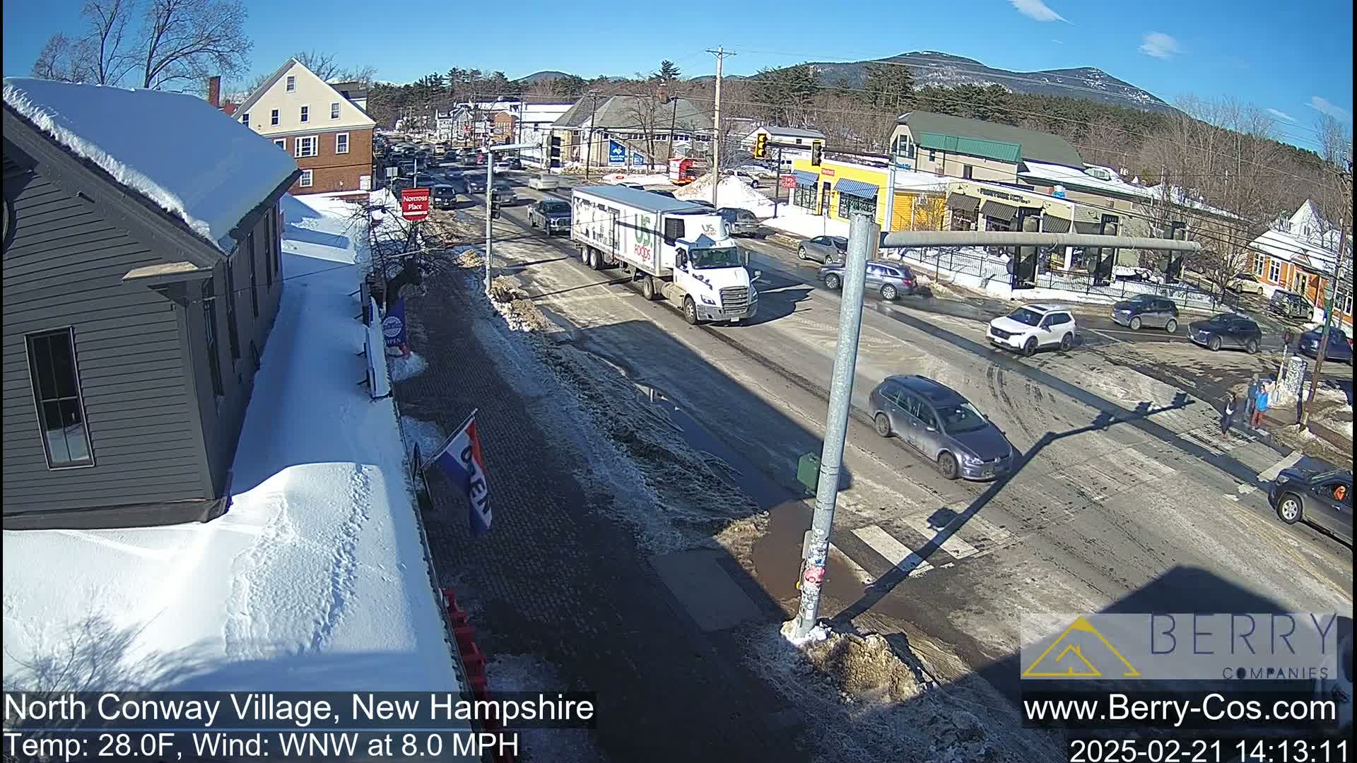 A sunny, snowy street scene shows cars and a large truck driving through a village intersection on a partly sunny day with a light wind.