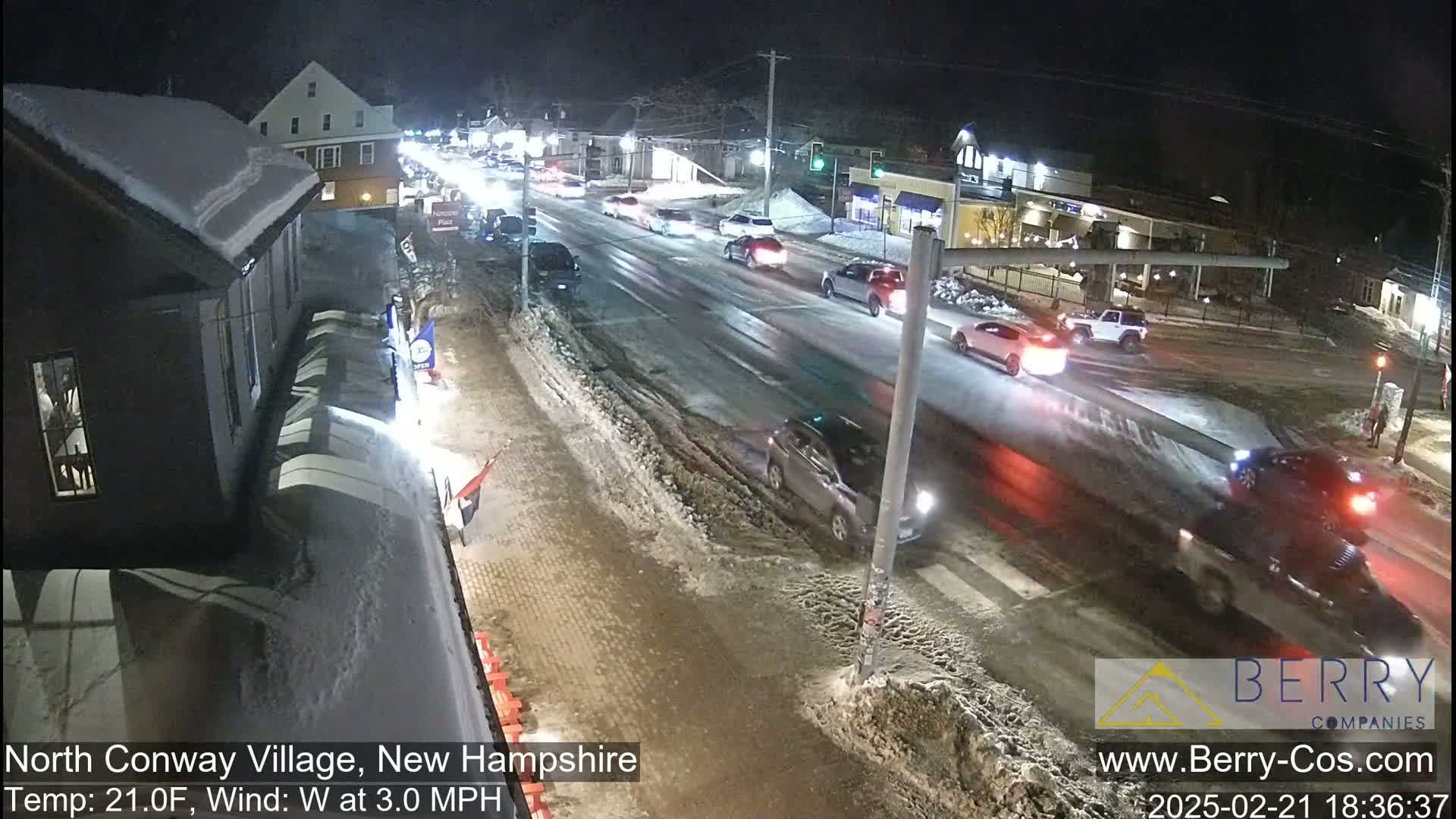 A snow-covered street scene at night shows several cars driving on a multi-lane road with buildings and businesses lining the sides.