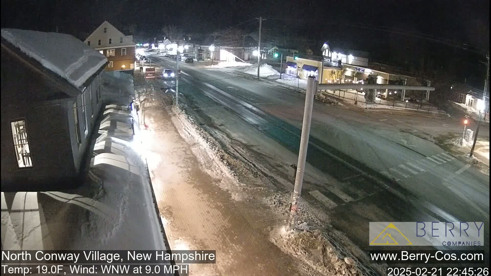 A nighttime street scene shows a snow-covered road with a few cars and buildings under a partly cloudy sky, with a temperature of 19 degrees Fahrenheit and a west-northwest wind at 9 miles per hour.