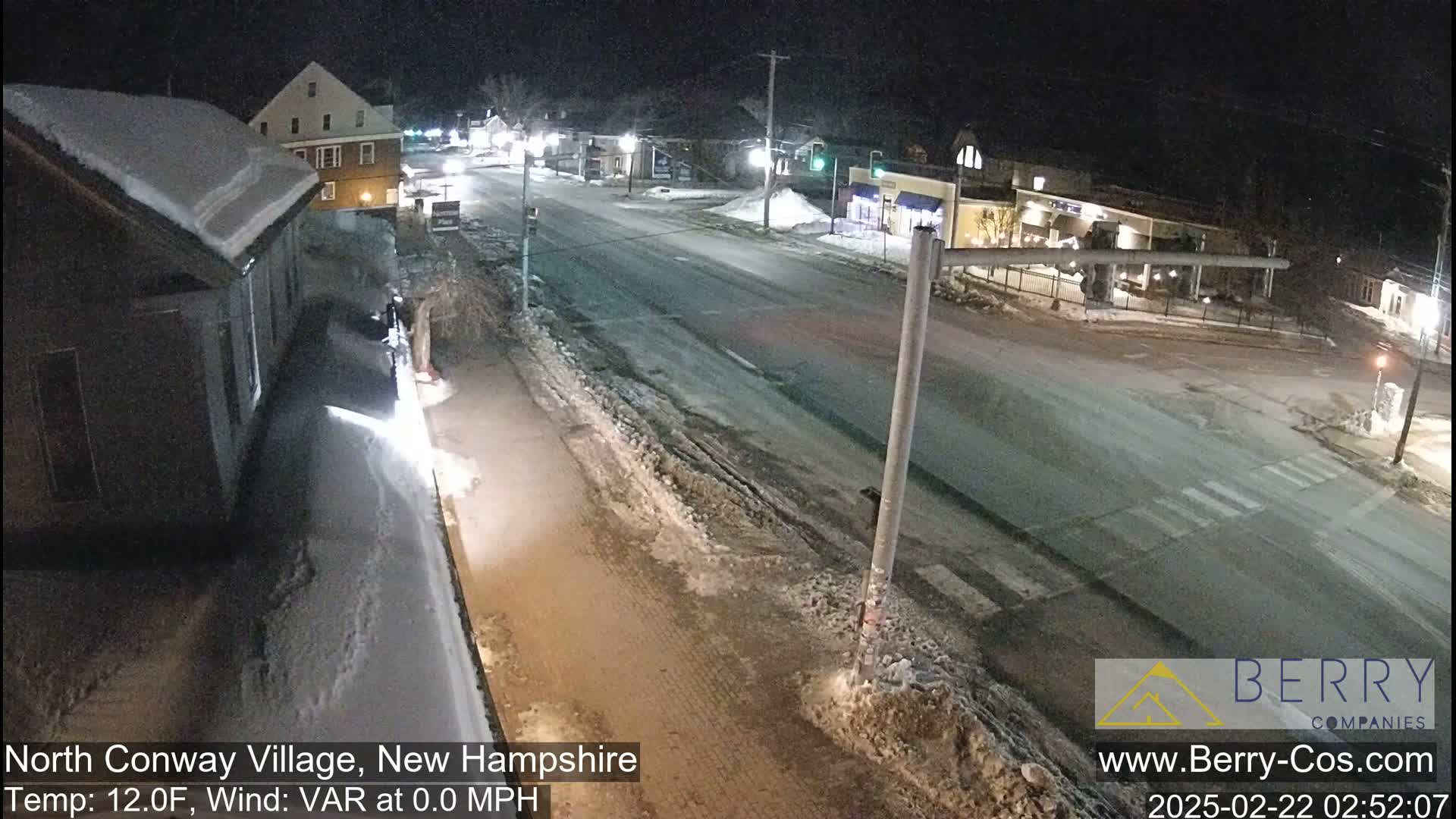 A snow-covered street scene at night shows a mostly empty road with buildings and streetlights visible in the distance under a clear sky.
