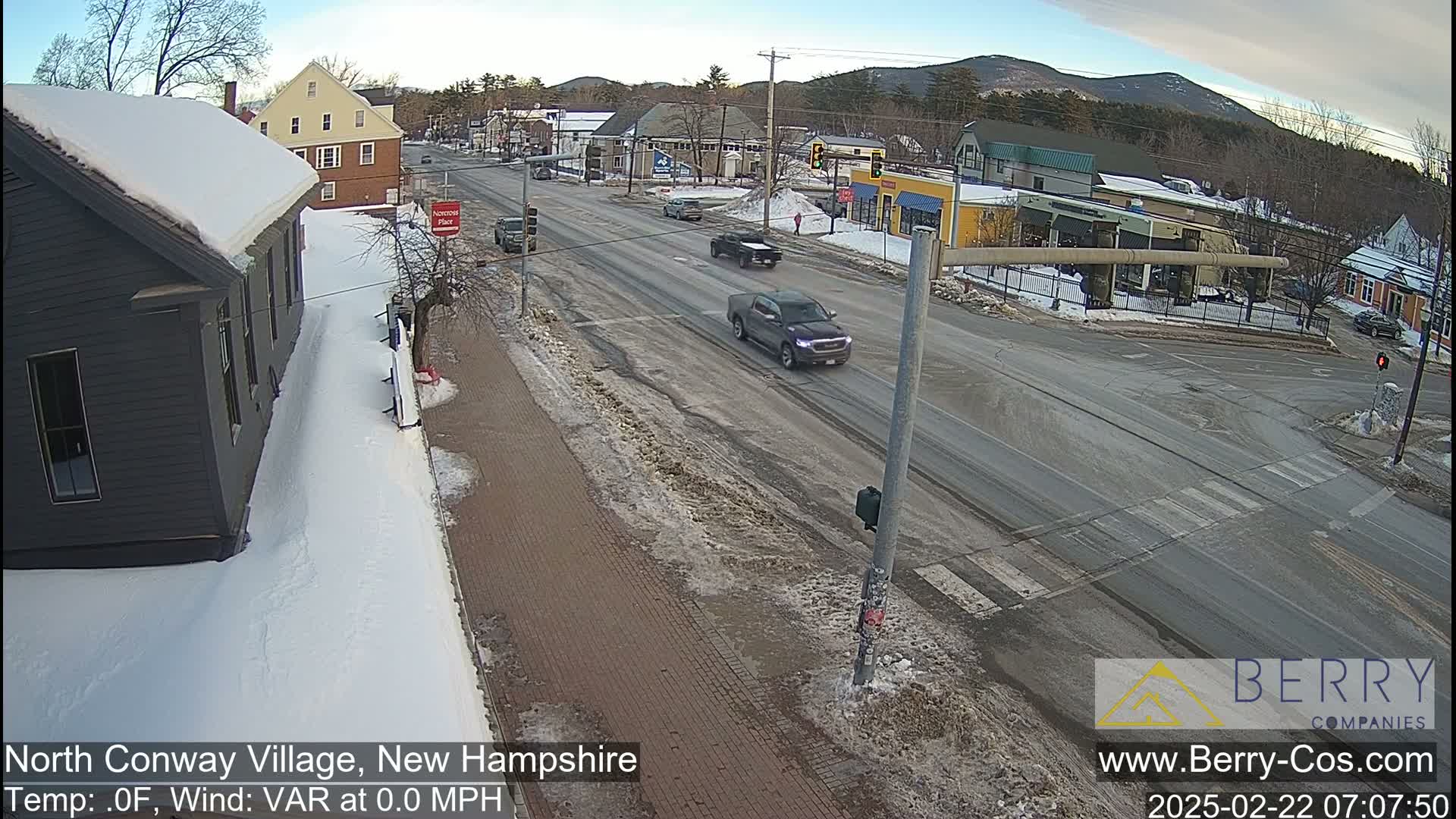 A snow-covered street scene shows several vehicles at an intersection in a village, with a background of low mountains under a clear sky.