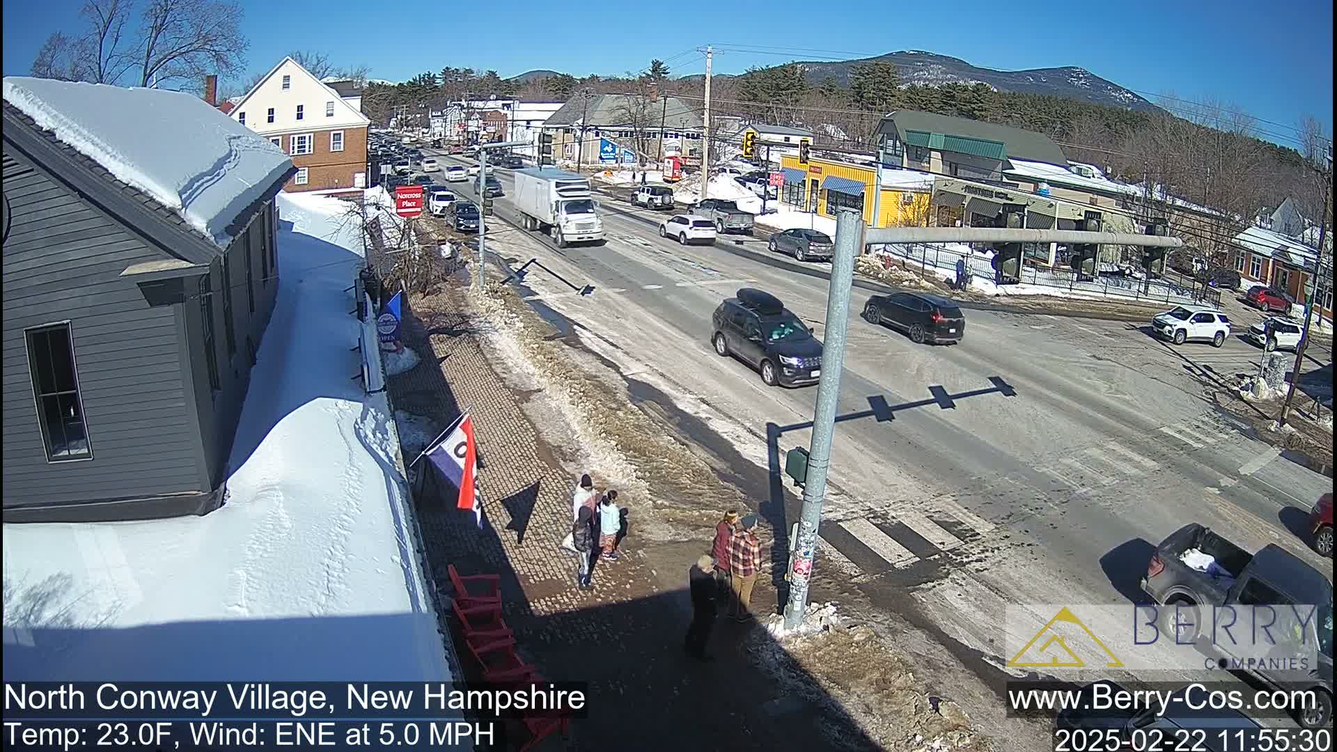 A sunny day reveals a snow-covered village street scene with several cars driving and pedestrians walking, some near a crosswalk.