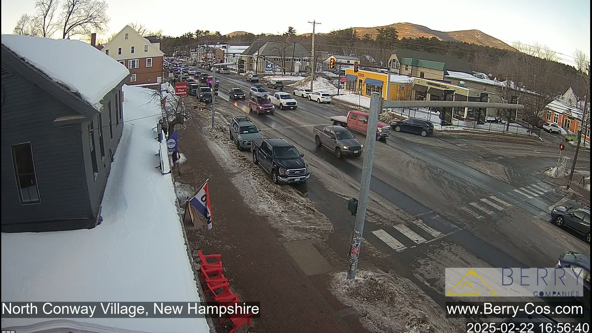 A snow-covered street scene shows several vehicles stopped and moving at an intersection in a village setting under a partly sunny sky.