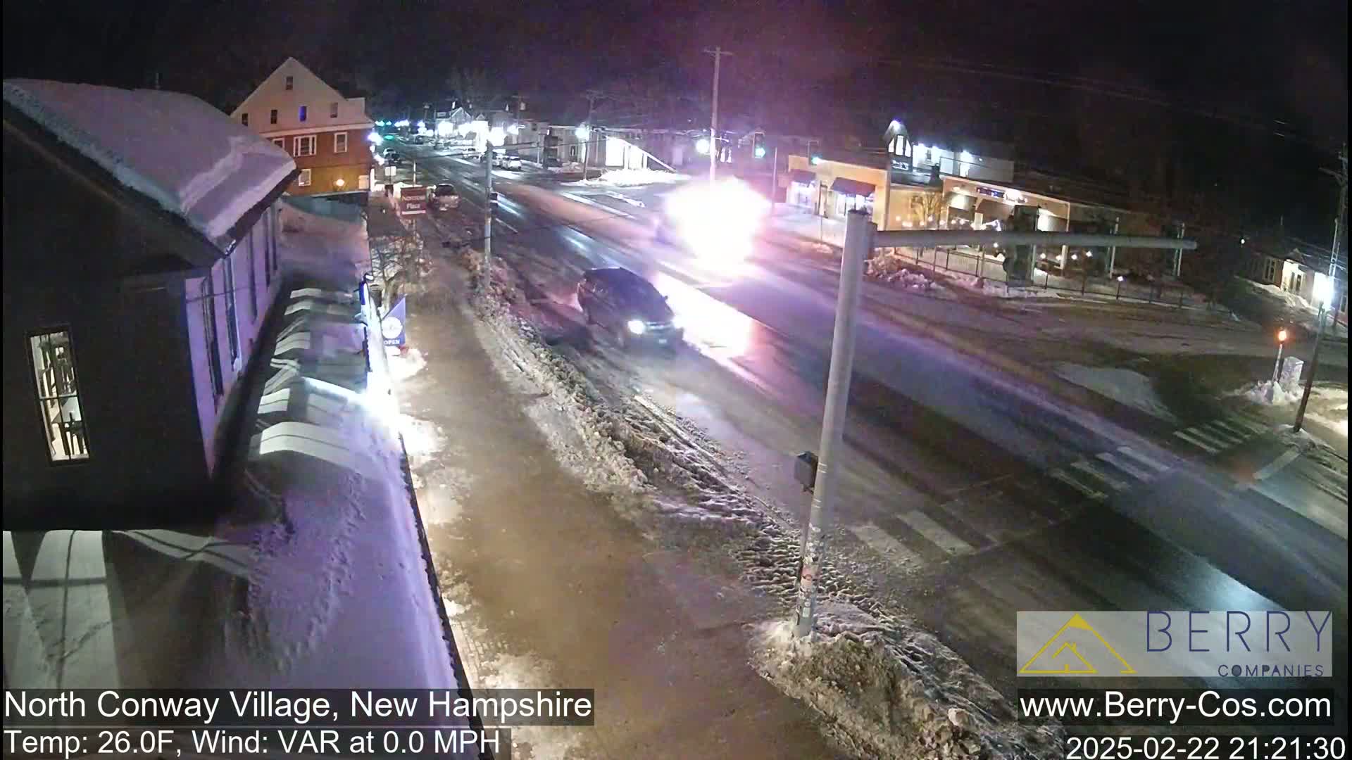 A nighttime scene of a snow-covered street with a vehicle driving past a bright flash, in a 26°F temperature with no wind.