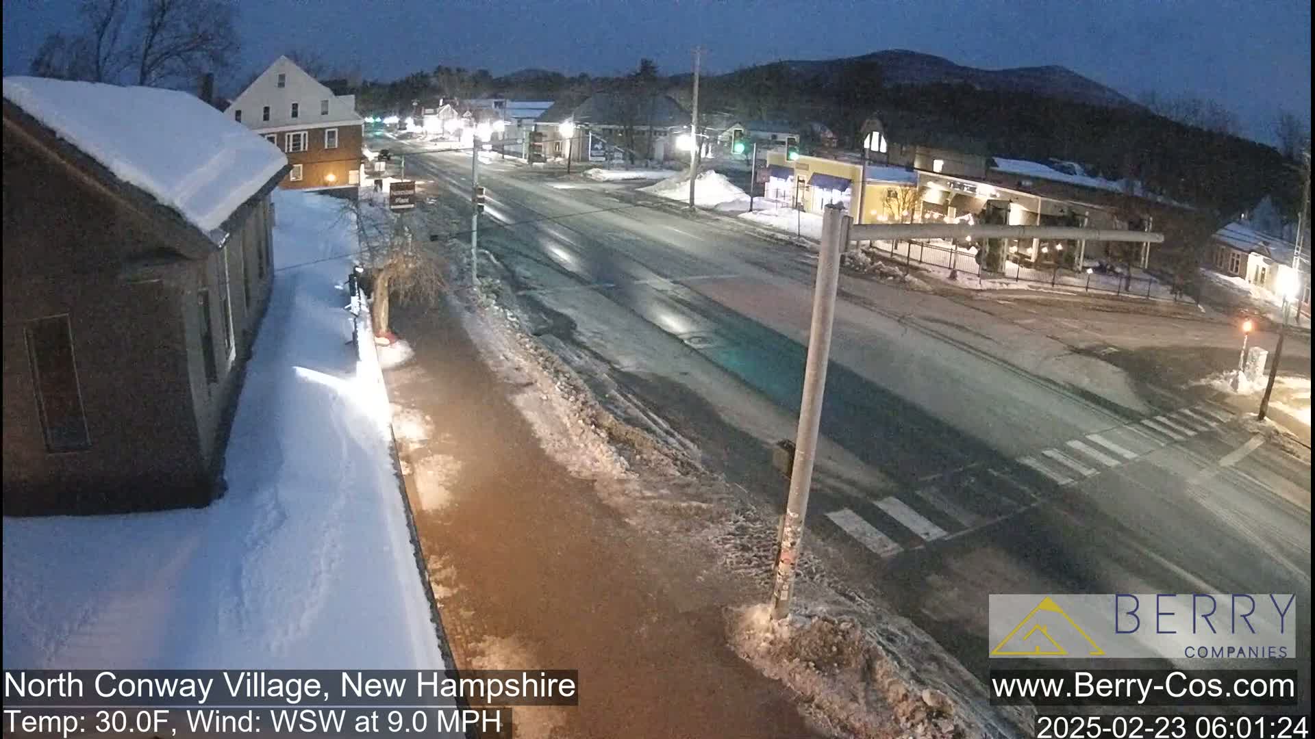A snow-covered street scene at night shows a road intersection with buildings and some pedestrian activity under a dark sky.