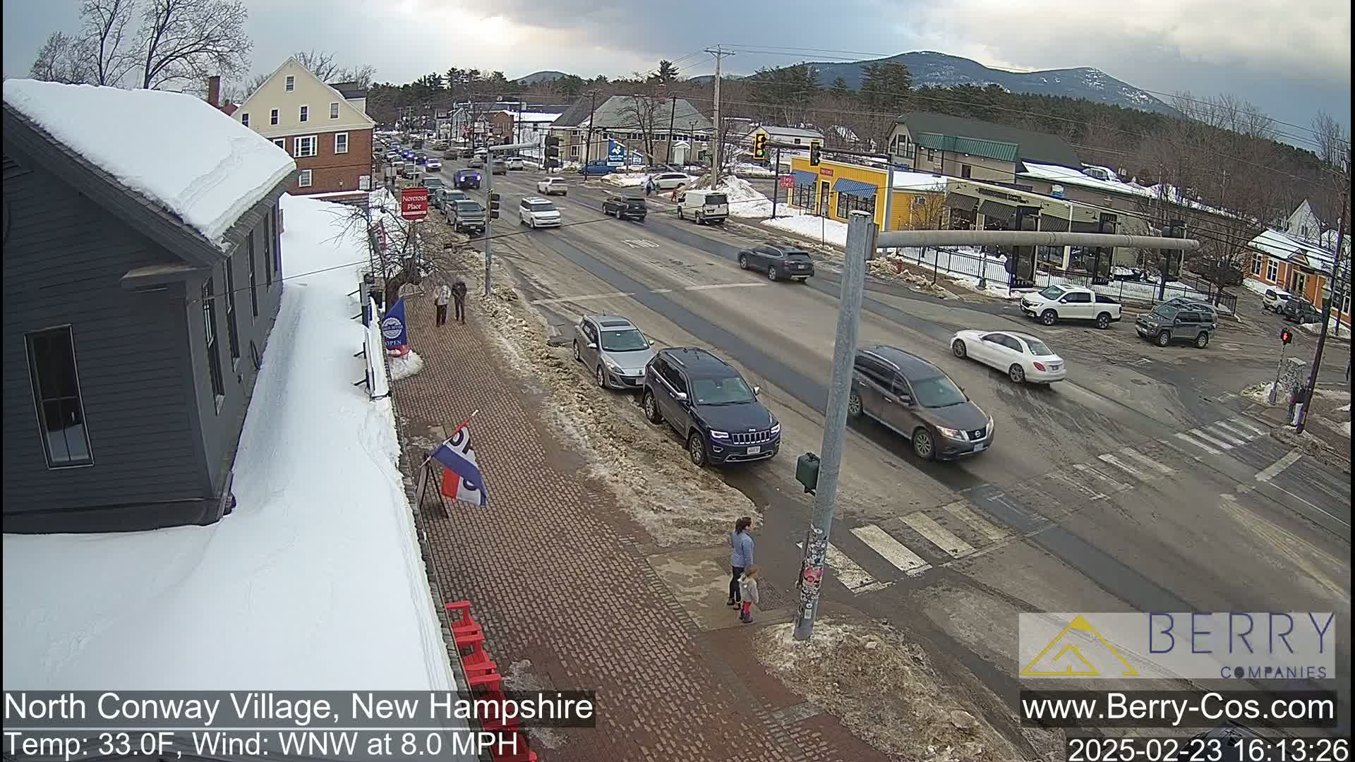 A snow-covered street scene in a village shows several cars parked and driving on a main road, with pedestrians walking on a brick sidewalk, under a cloudy sky.