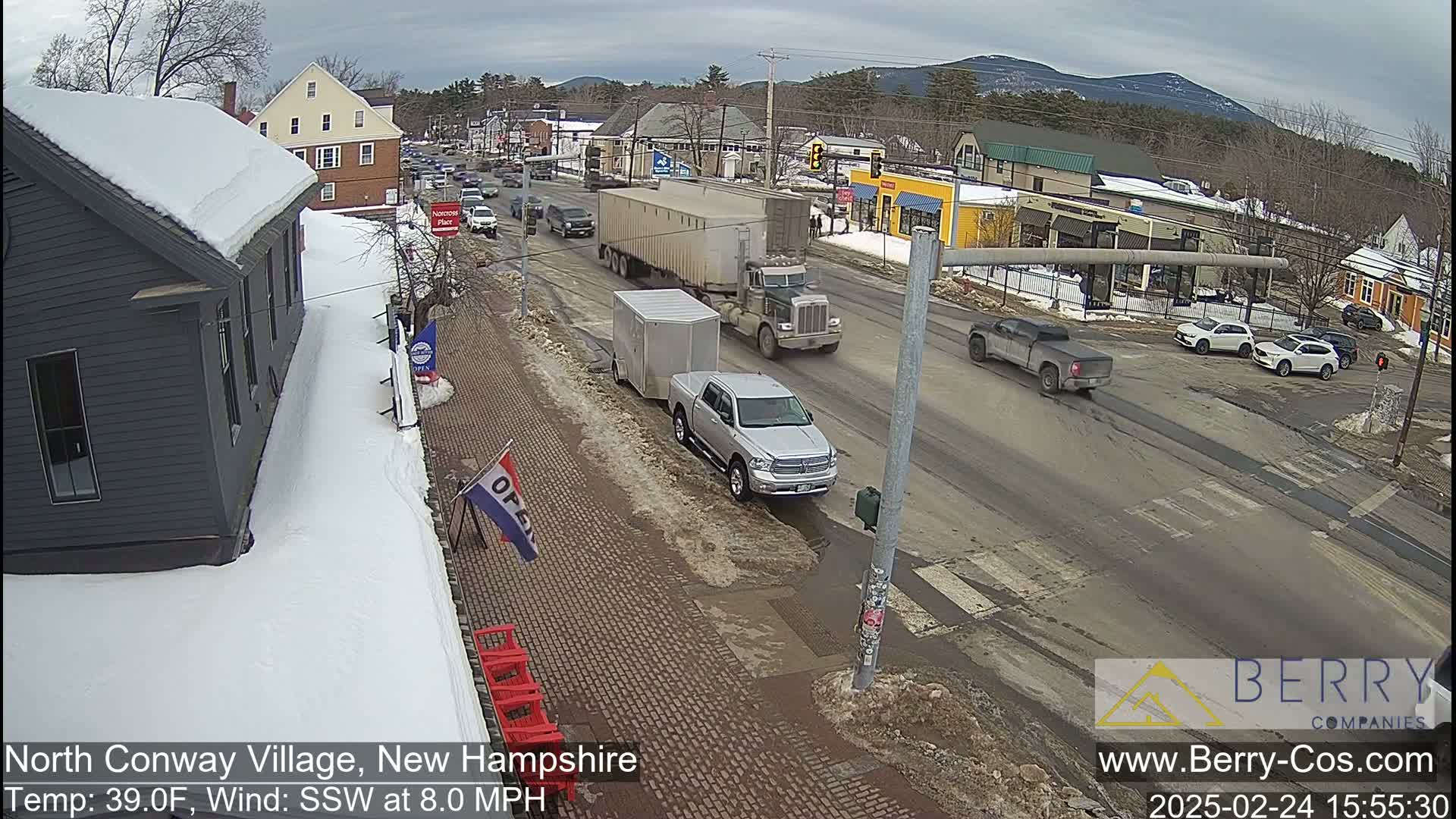 A snow-covered street scene shows several vehicles, including trucks and cars, driving on a road that passes in front of small businesses in a village under an overcast sky.