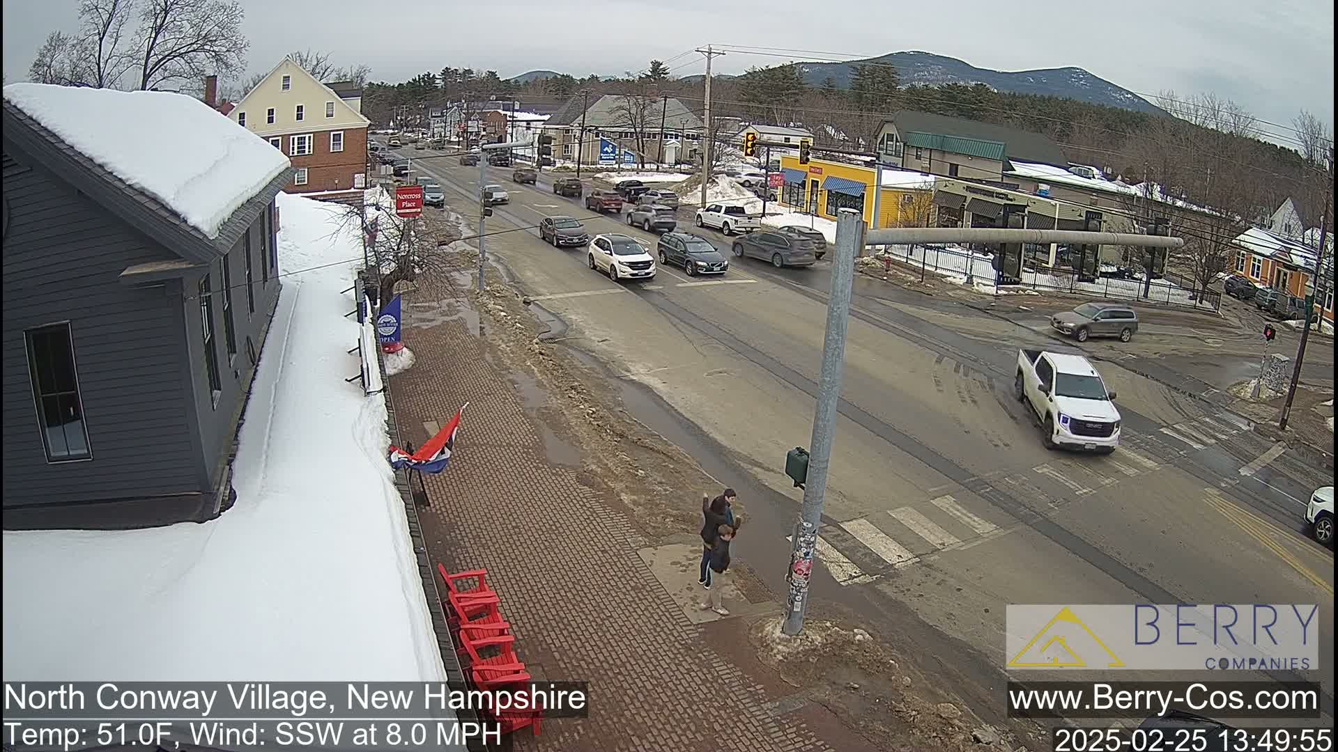 A partly sunny, snowy street scene shows several cars driving on a road past shops and buildings, with two people standing on a sidewalk near red chairs.