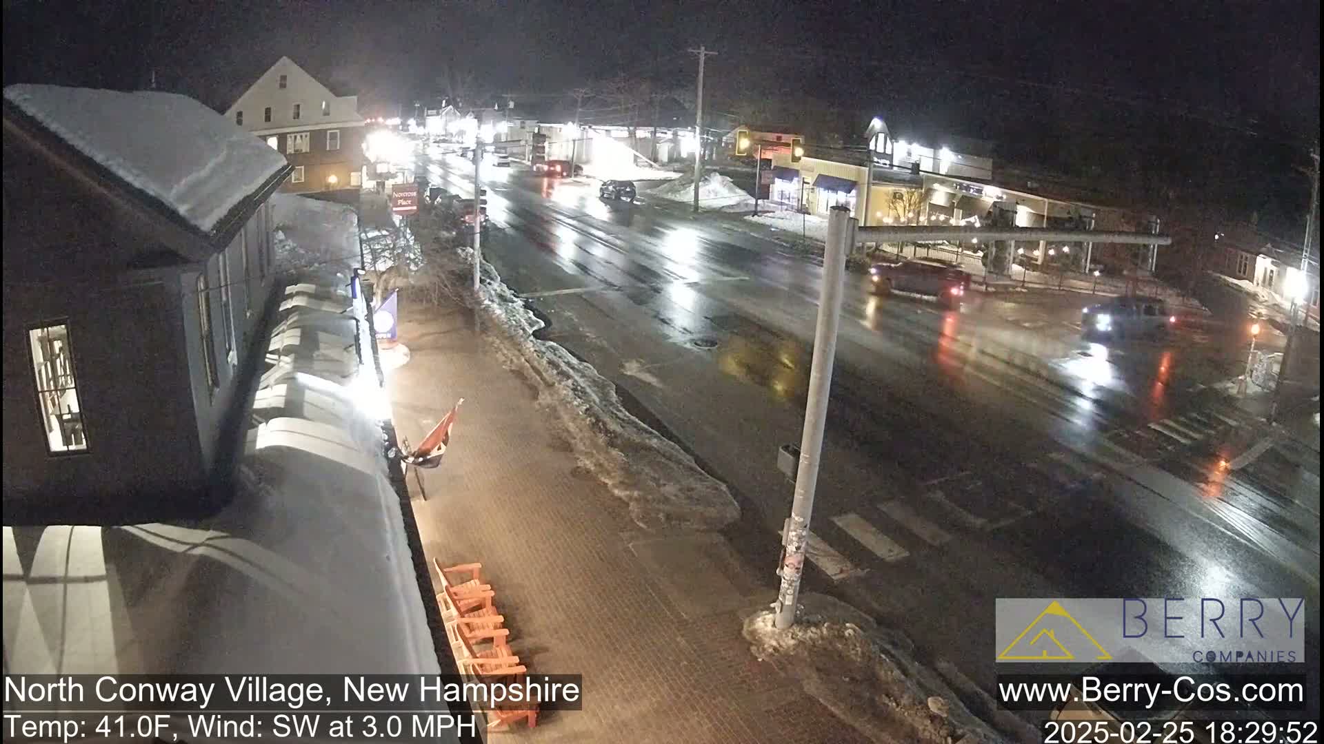 A wet, nighttime street scene shows a few cars at an intersection in a small town, with snow and ice visible on the sidewalks and some buildings in the background.
