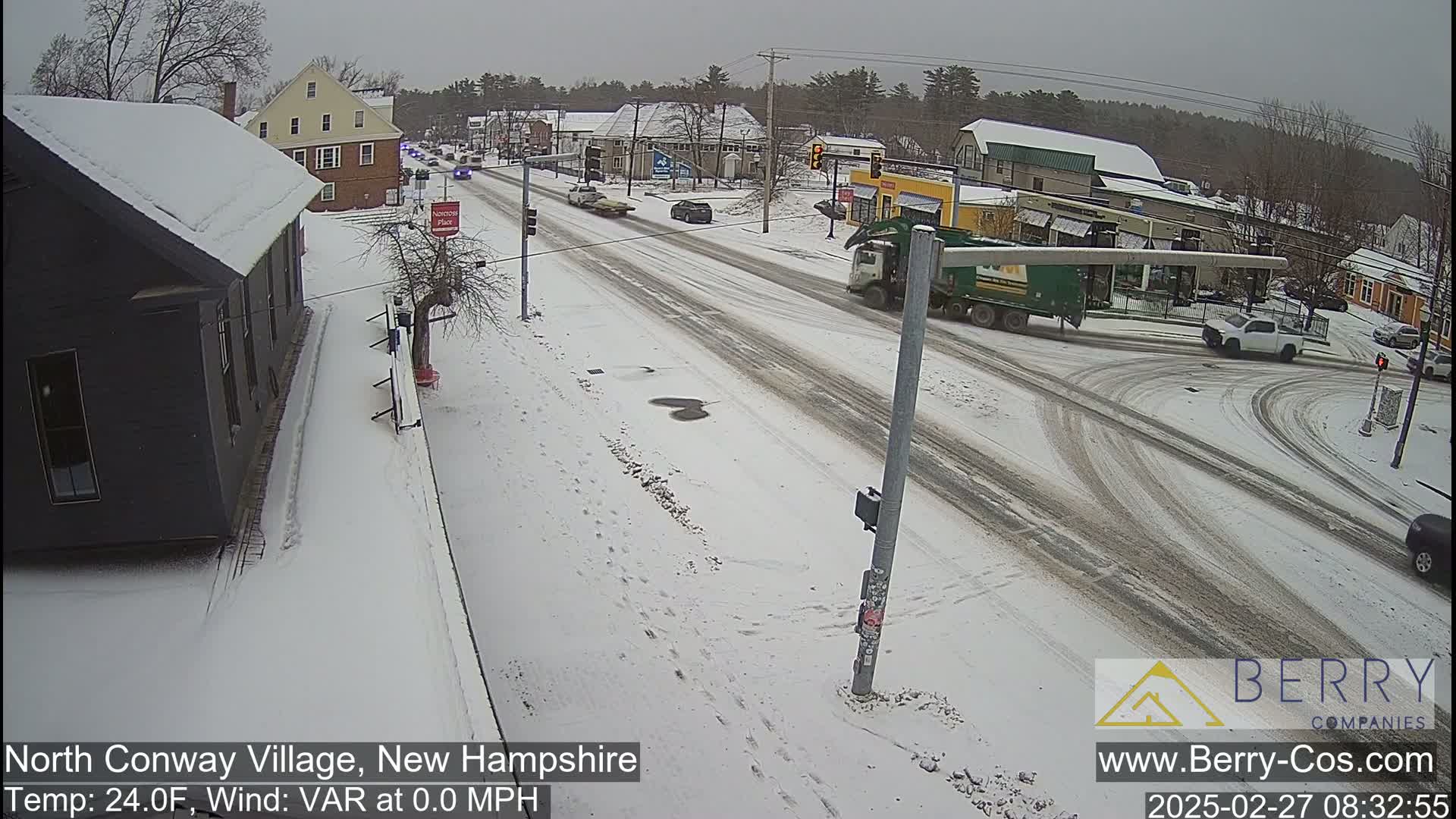 A snow-covered street scene shows several cars and a garbage truck at an intersection, with buildings and trees lining the street in a light snowfall.