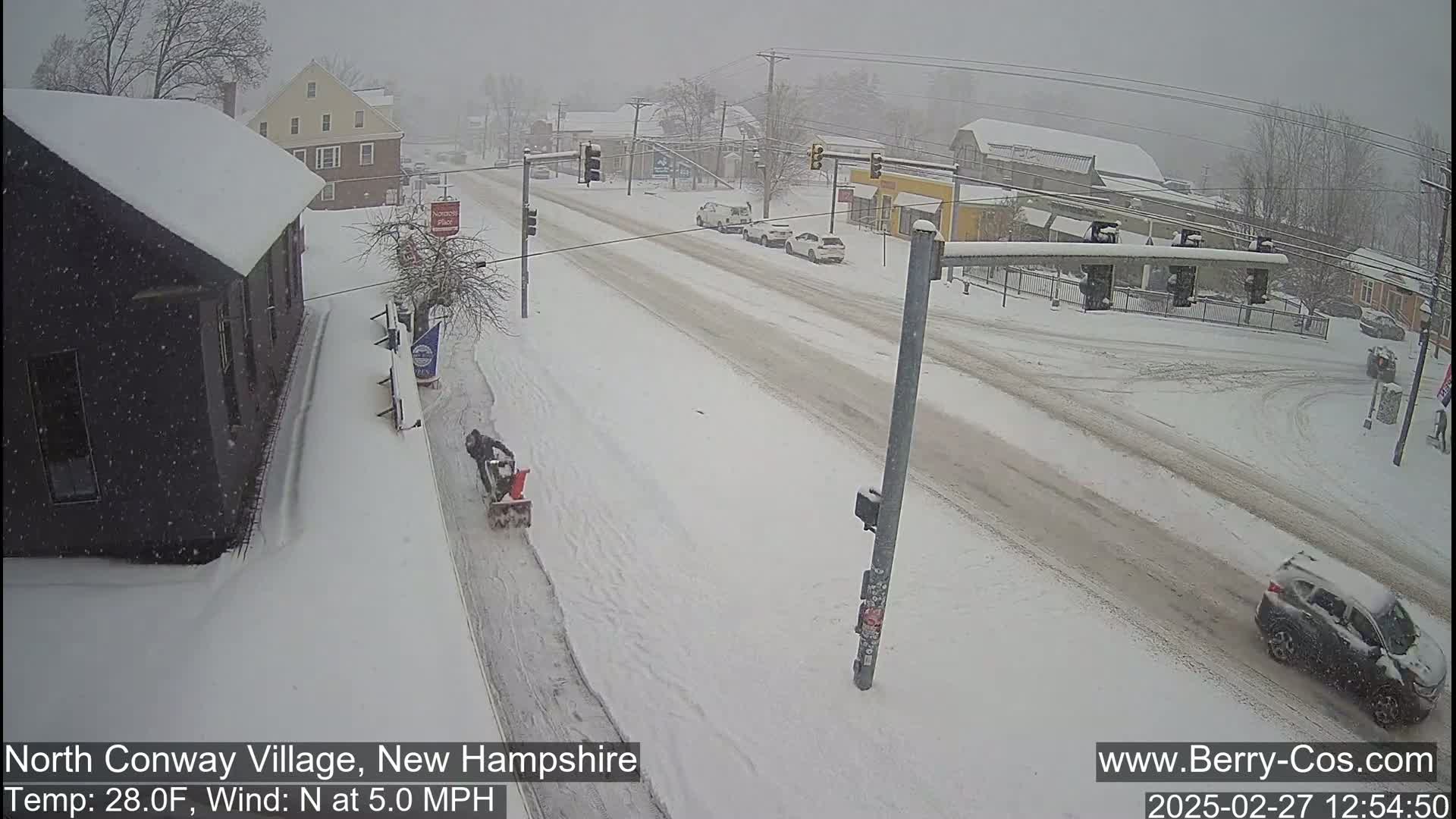 A snow-covered street scene shows a person using a snow blower on a sidewalk, several cars driving on a road, and buildings covered in snow during a snowfall.