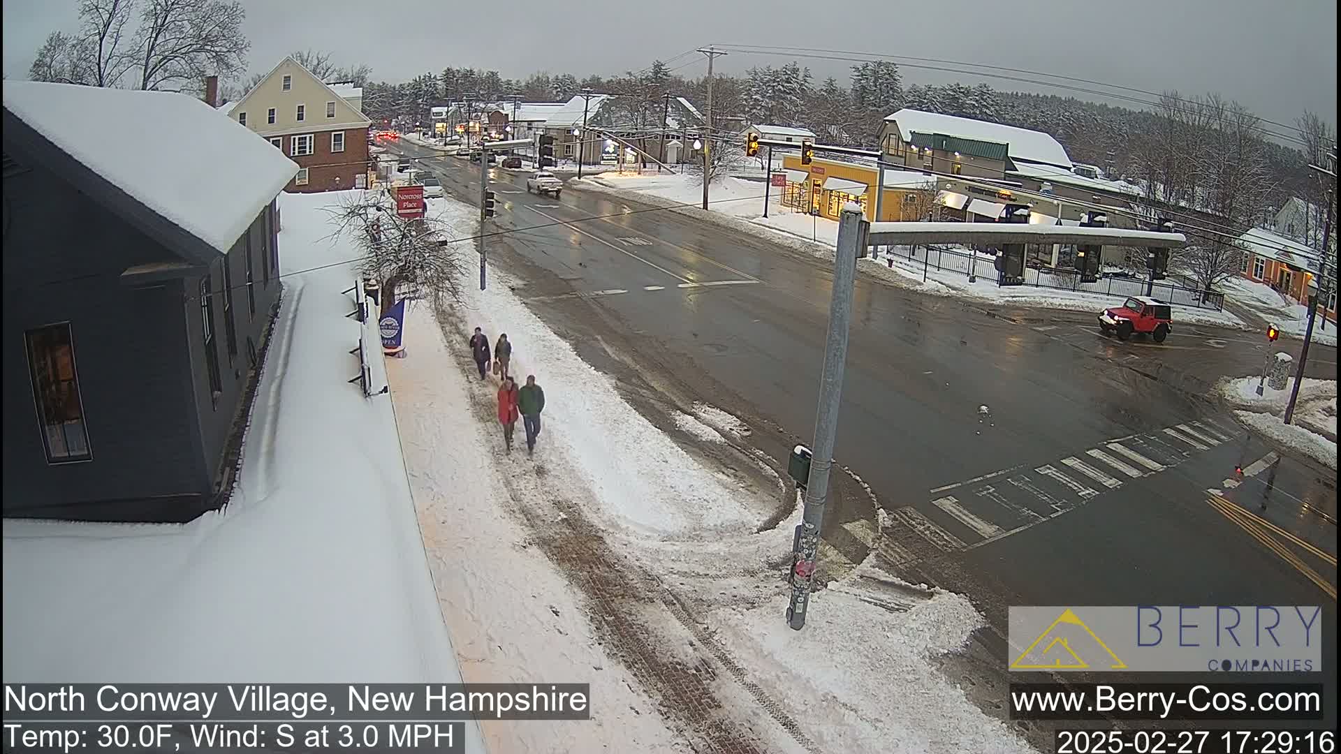 A snow-covered street scene shows several pedestrians walking on a sidewalk, cars driving on a wet road, and buildings in the background during a light snowfall.