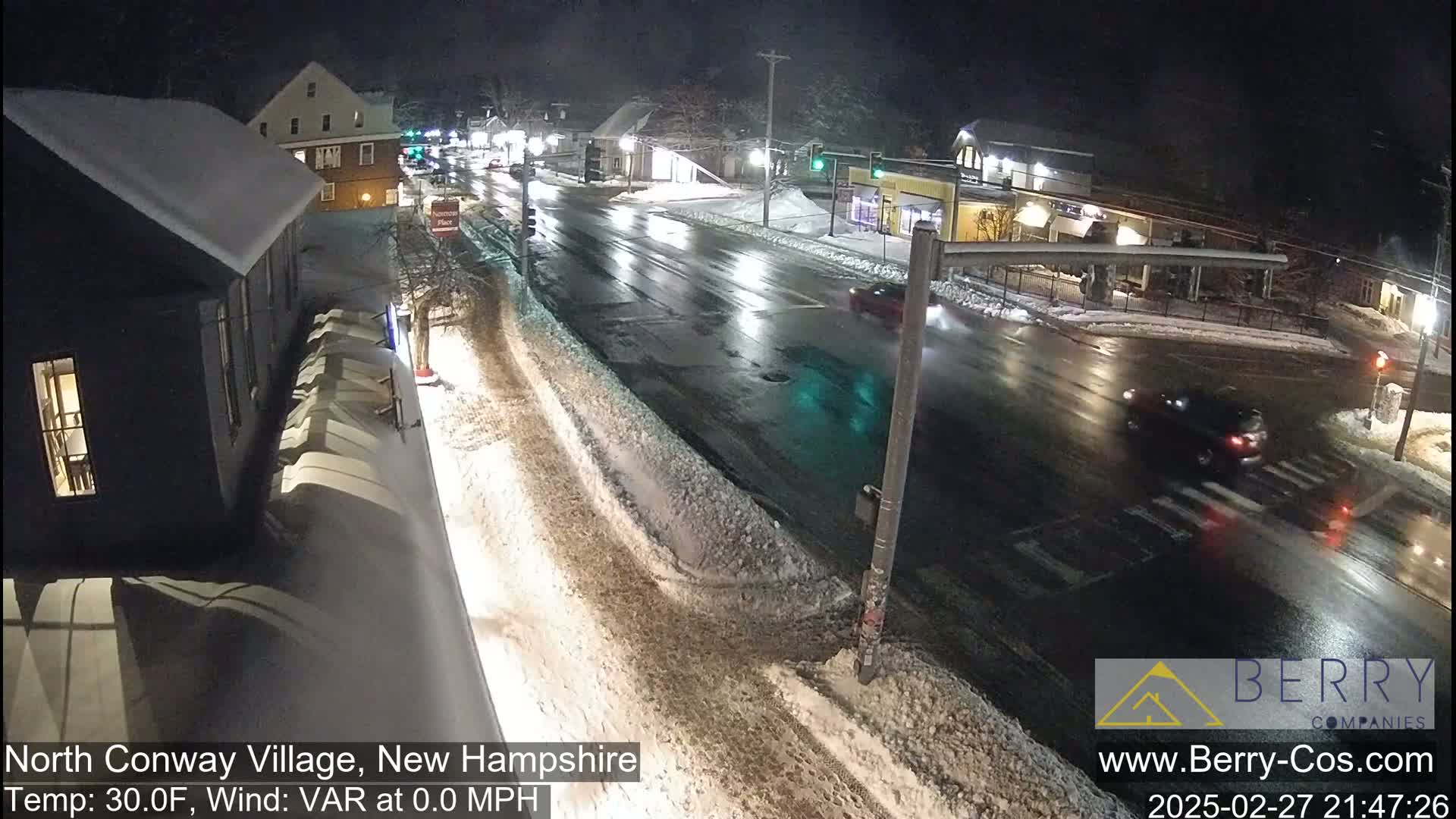 A snow-covered street scene at night shows a few cars driving on a wet road, with buildings and streetlights visible in the background.