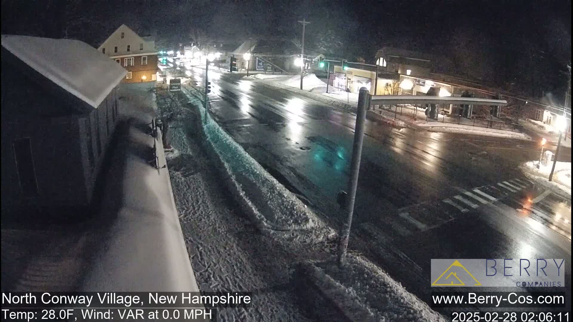 A snow-covered street scene at night shows a wet road with some cars and buildings under a dark sky.