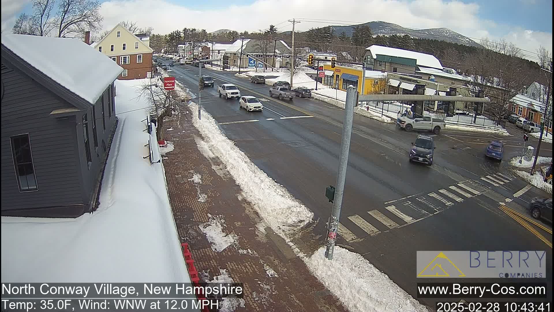 A snow-covered street scene shows several vehicles at an intersection in a town, with mountains in the background under a partly cloudy sky.