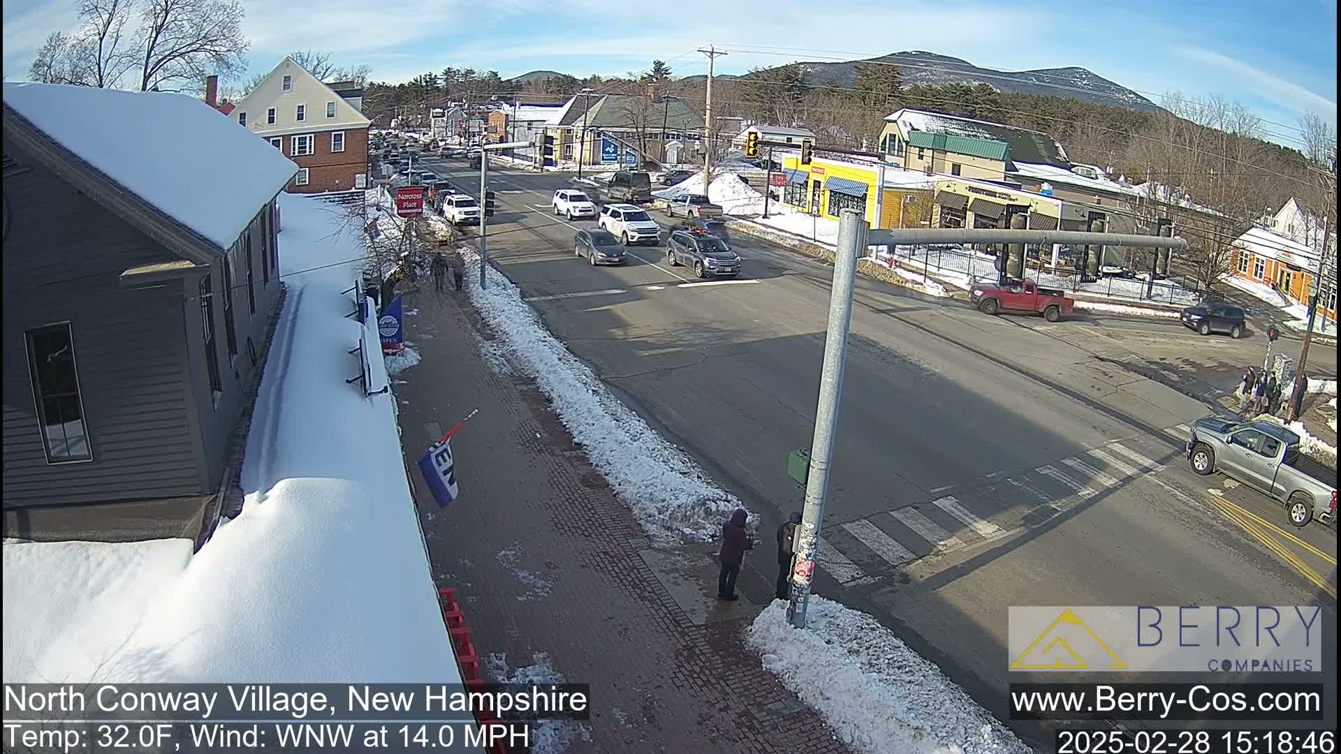 A sunny, snow-covered street scene shows several vehicles at an intersection, with pedestrians walking on the sidewalk and snow banks lining the road.