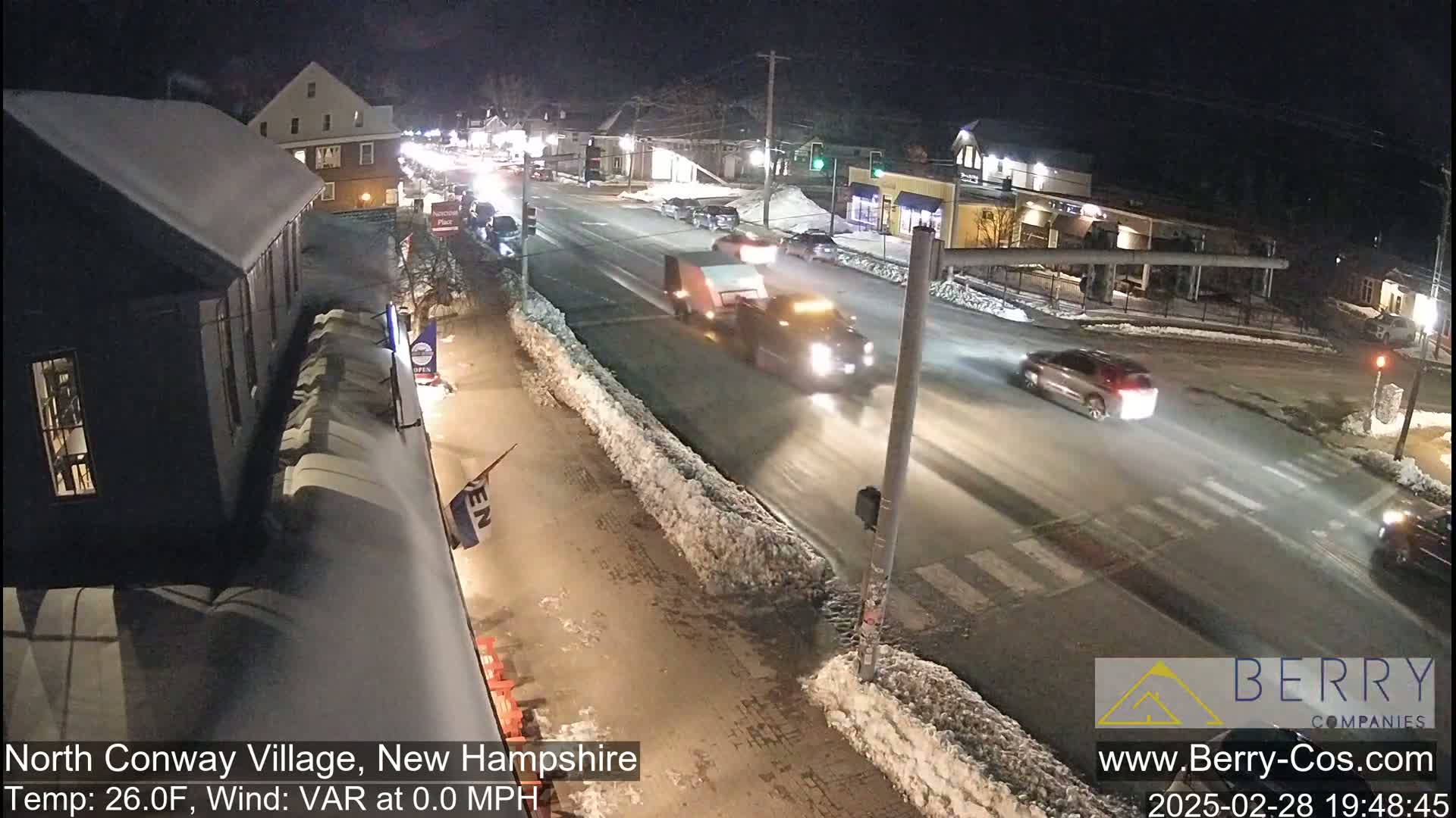 A snow-covered street at night with several vehicles driving on a road, and buildings are visible in the background under dark, clear skies.