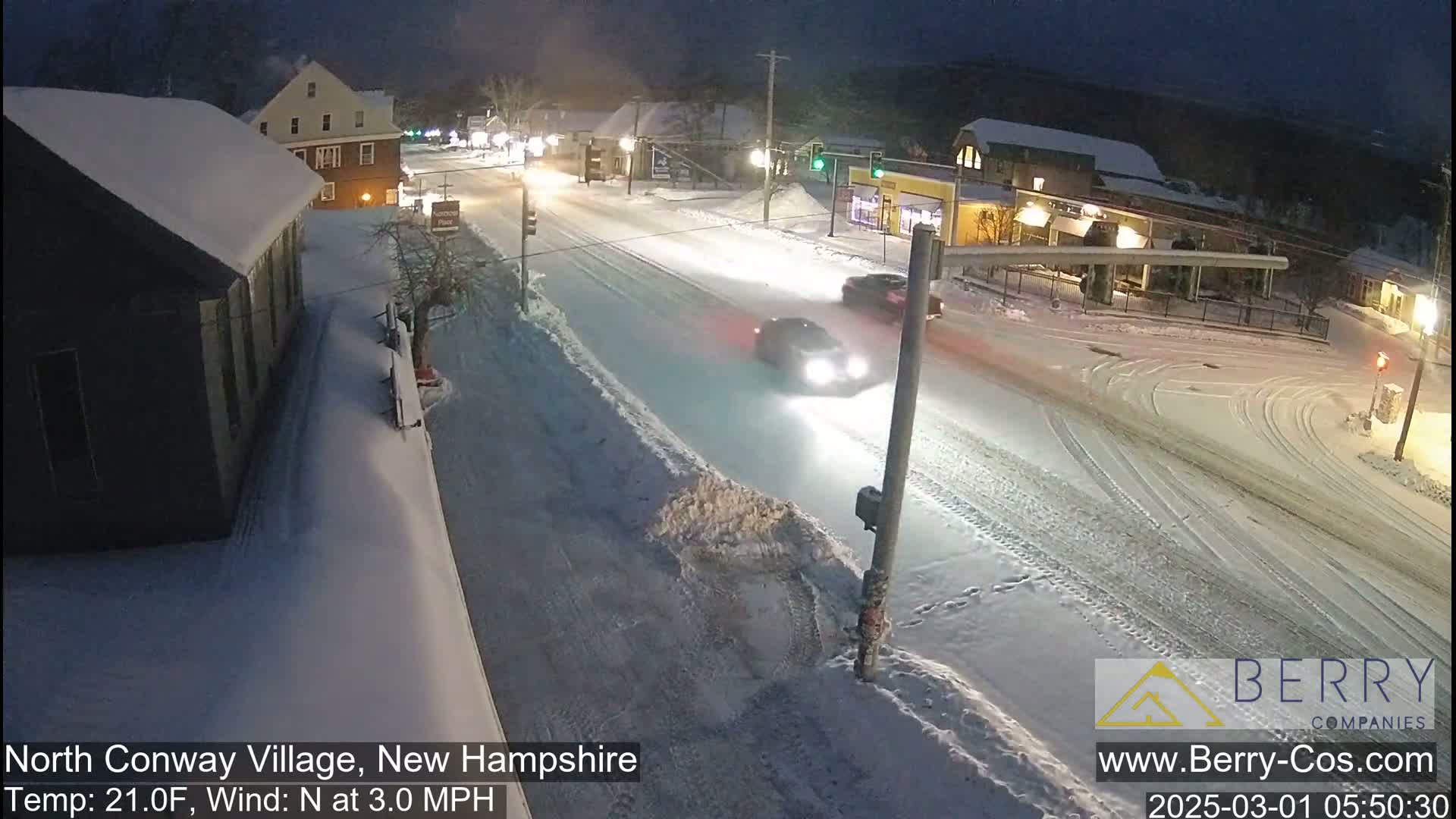 A snow-covered street scene at night shows several cars driving through a town, with buildings on either side of the road.