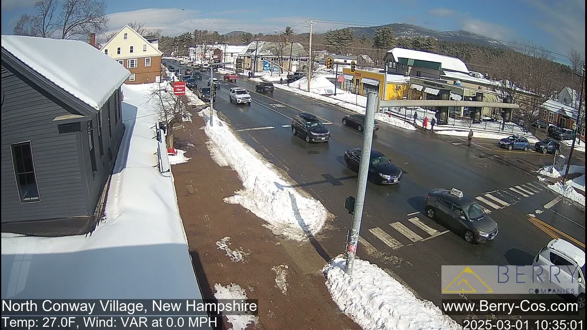 A sunny winter day shows a snow-covered street scene with several cars driving through a village intersection.