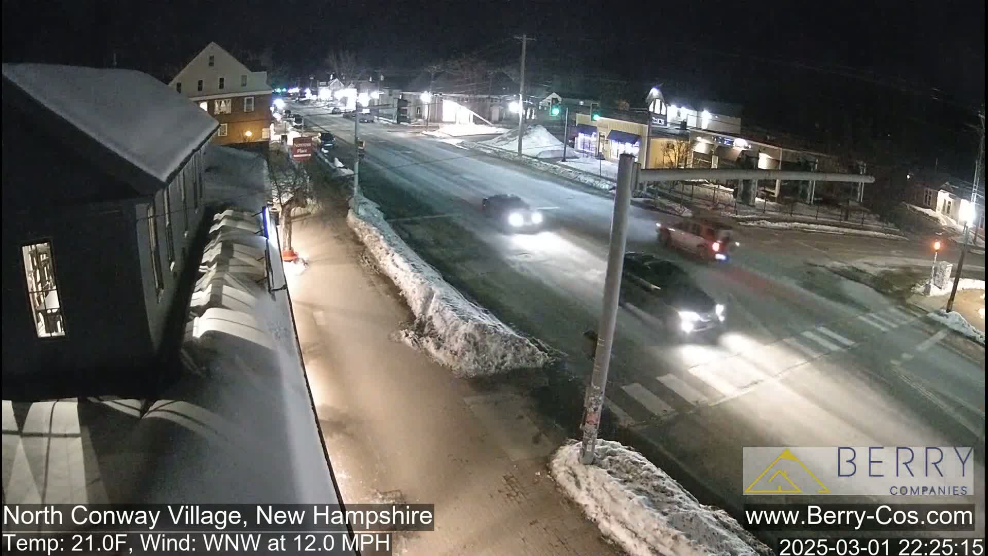 A snow-covered street scene at night shows several cars driving through a town intersection under a clear sky with a 21°F temperature and a 12 mph wind.