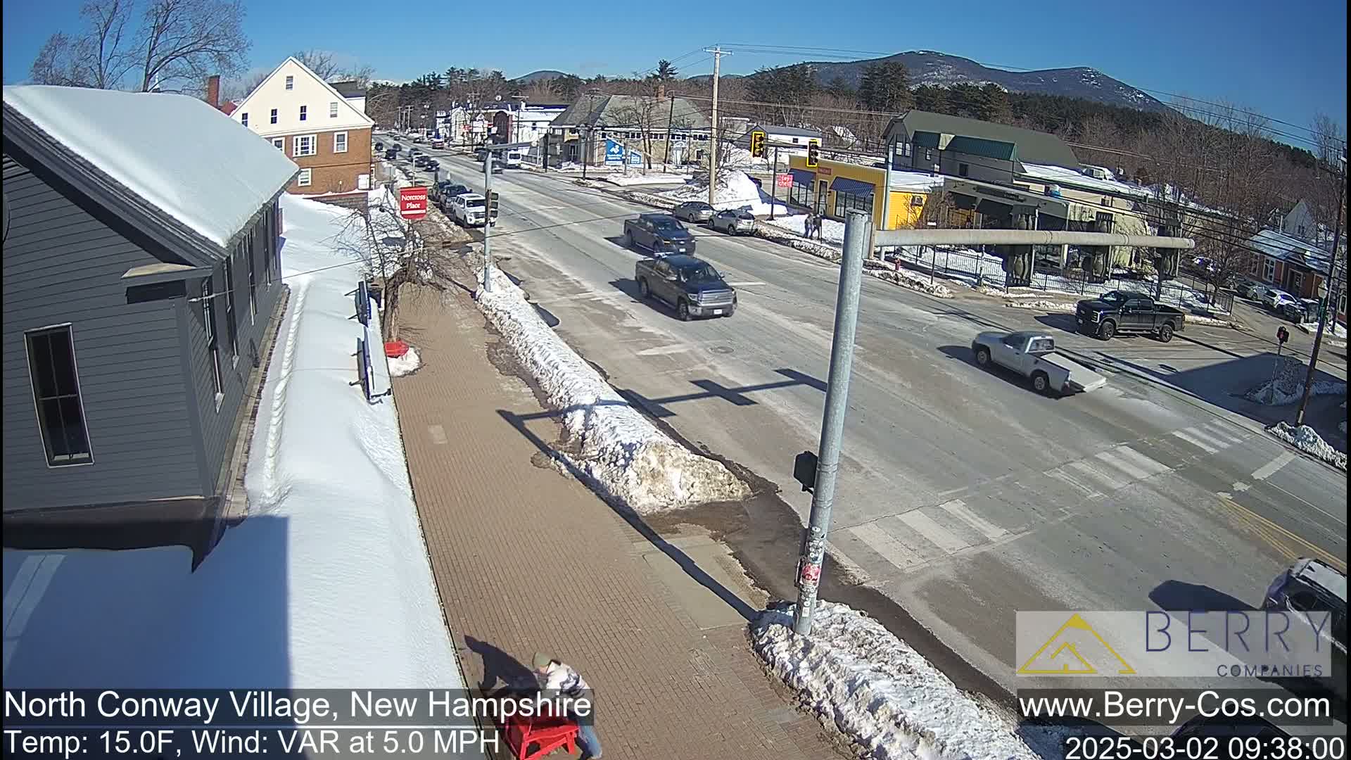 A sunny day with snow on the ground and sidewalks shows a street scene with several vehicles, some snow-covered buildings, and a person near a red object.