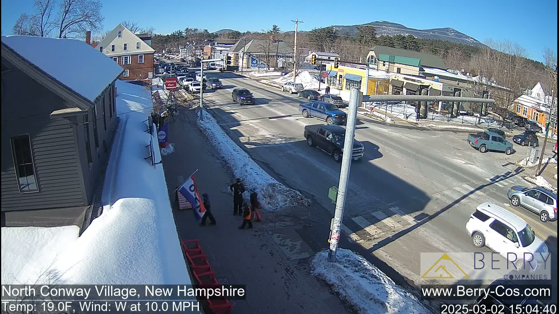 A sunny, snow-covered street scene shows several vehicles at an intersection, with a few pedestrians near a building on the left; a mountain is visible in the background.