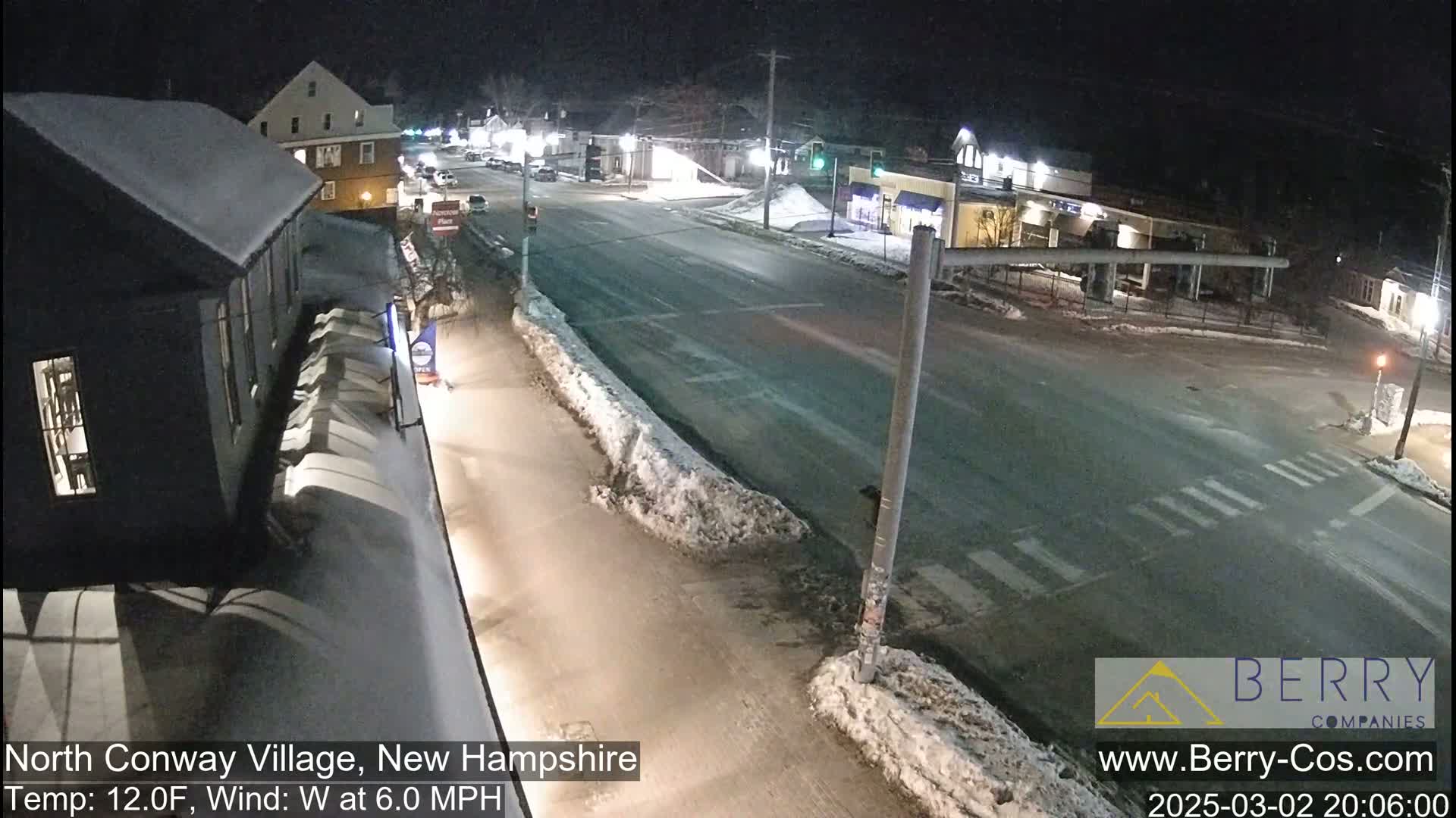 A snow-covered street intersection at night, with a few buildings and cars visible, under a clear sky.
