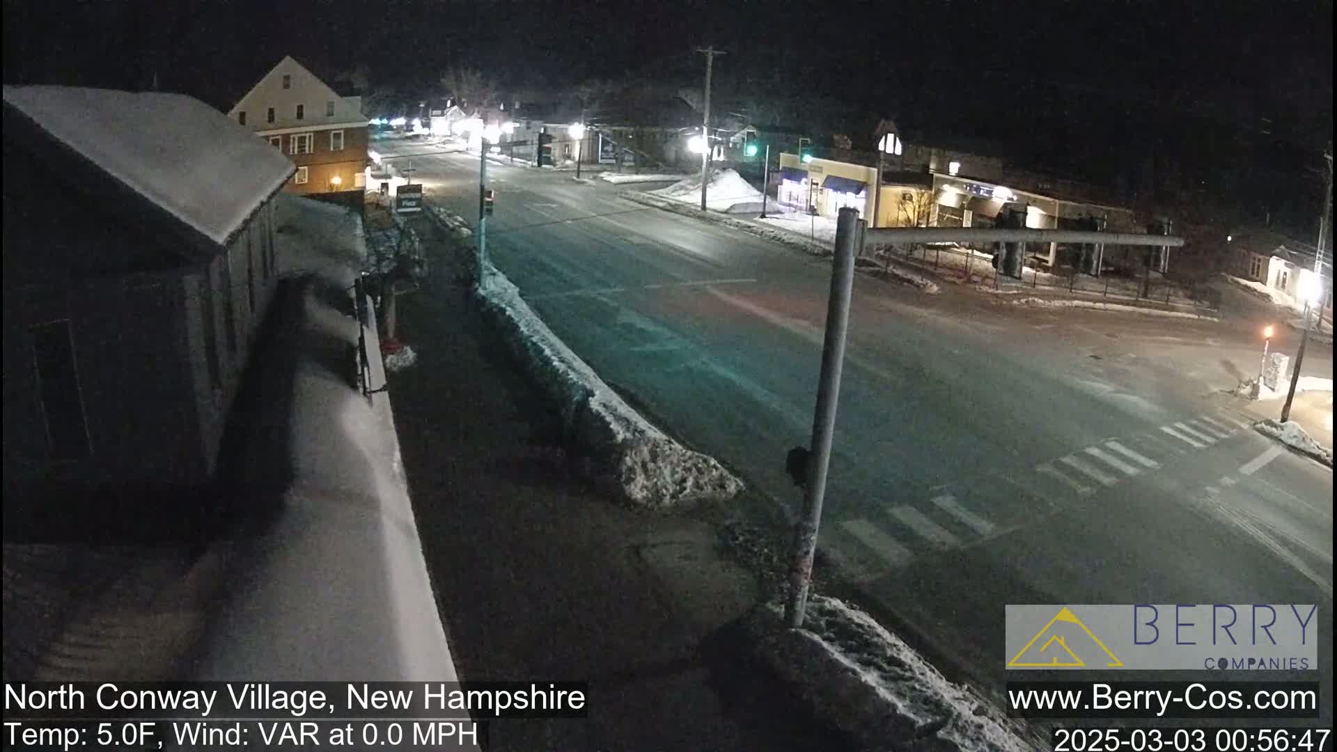 A snow-covered street intersection at night, with a few buildings and some cars visible.